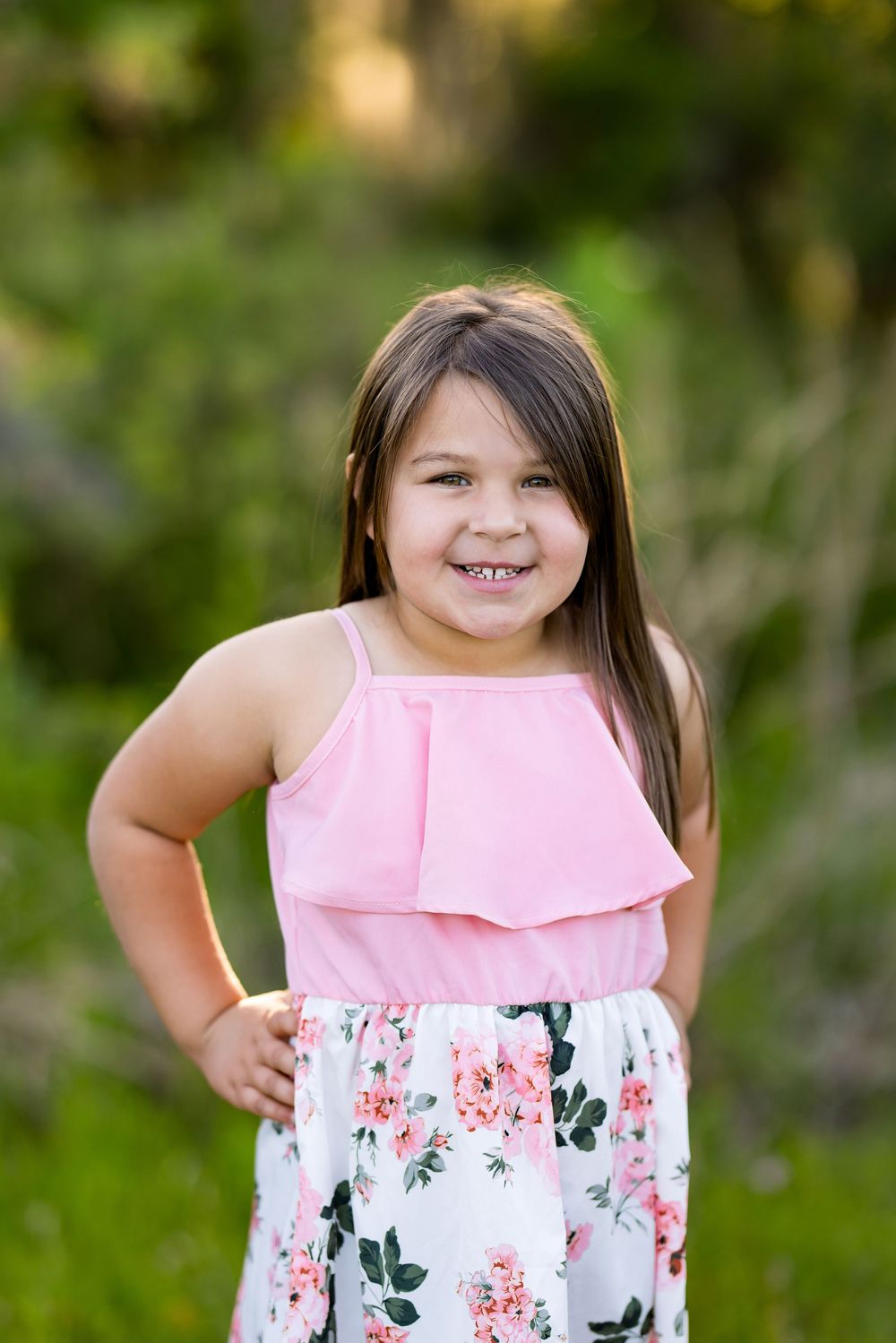 A young child wearing a pink sleeveless top and floral skirt stands outdoors in a natural setting during golden hour.