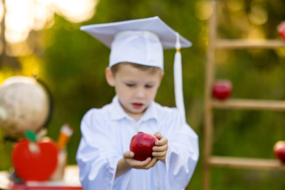 Young child in a white shirt carefully examines a red apple in an outdoor garden setting with soft bokeh background.