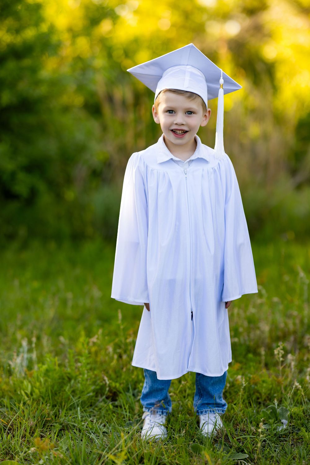 A graduate in a white cap and gown stands in grass during a sunny outdoor photo session.