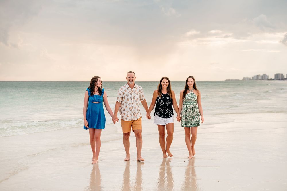 Four people walk hand in hand along a pristine white beach at sunset with the ocean and cityscape in the background.