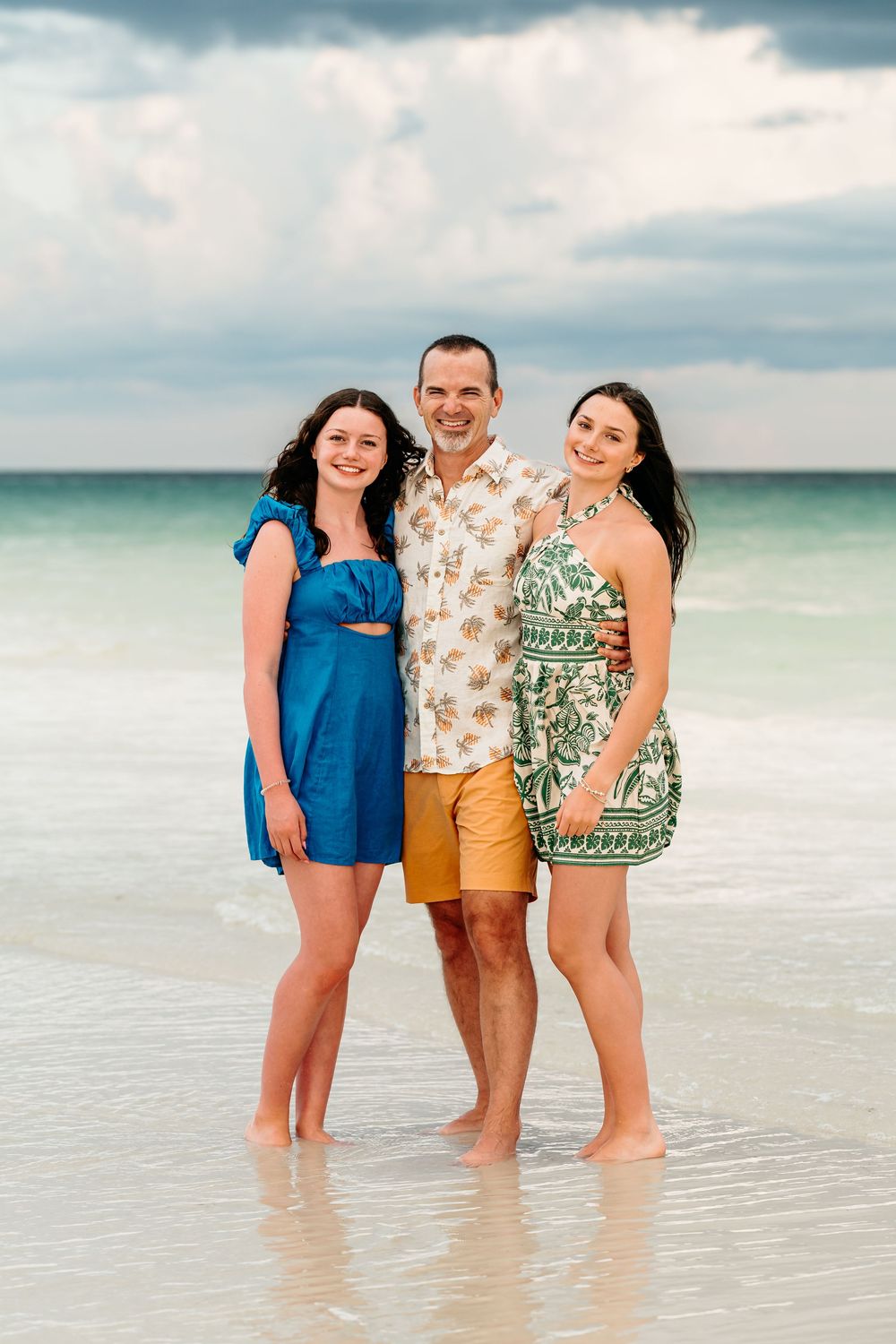 Three people in summer clothing standing together on a beach with turquoise water and cloudy sky in the background.
