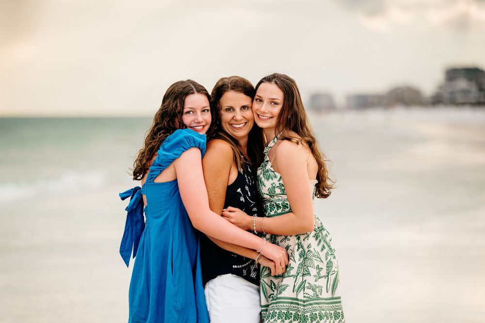 Three women in casual summer outfits smile and embrace on a beach at sunset while taking photos together.