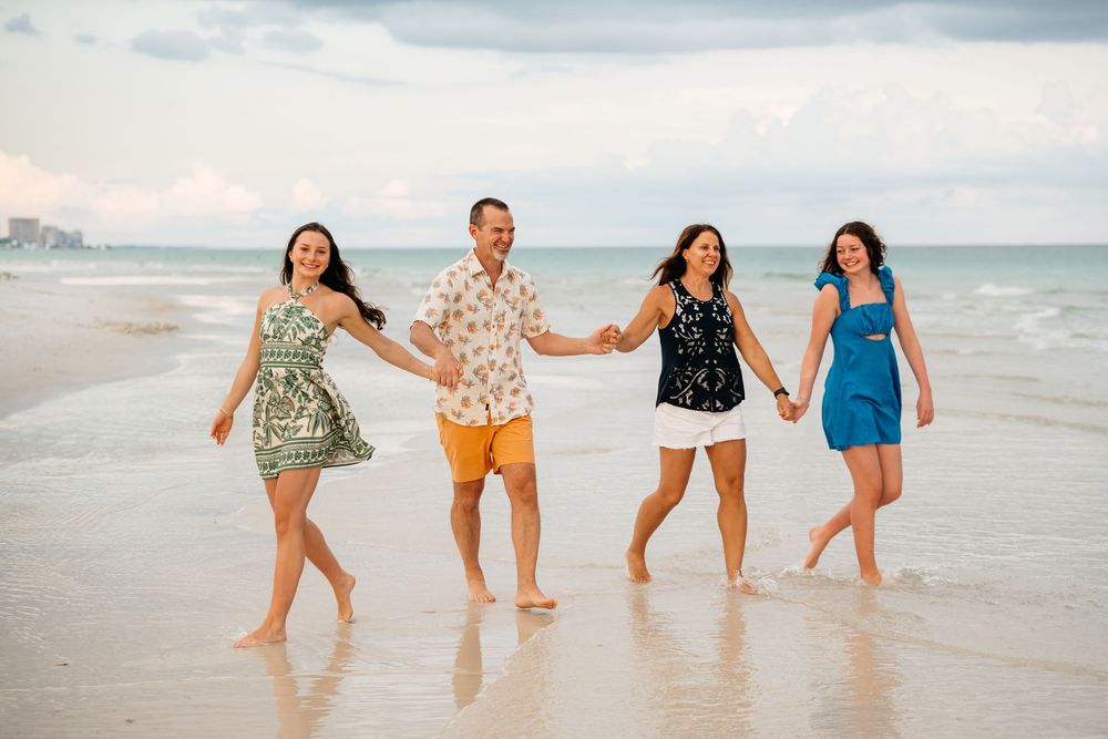 Four friends in summer outfits holding hands while walking and laughing together on a cloudy beach.