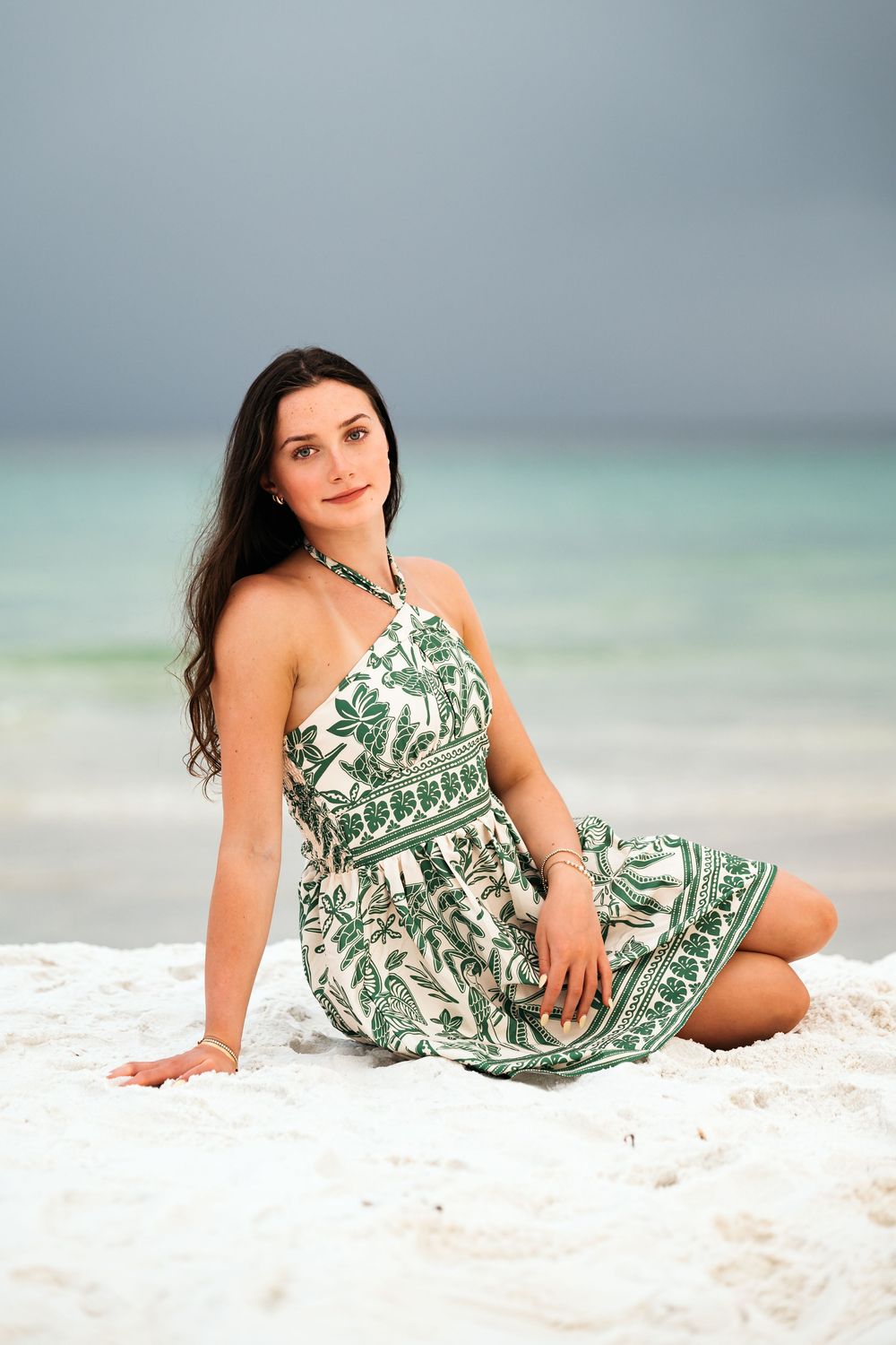 Woman in tropical green and white palm print dress poses gracefully on white sandy beach with turquoise ocean background.