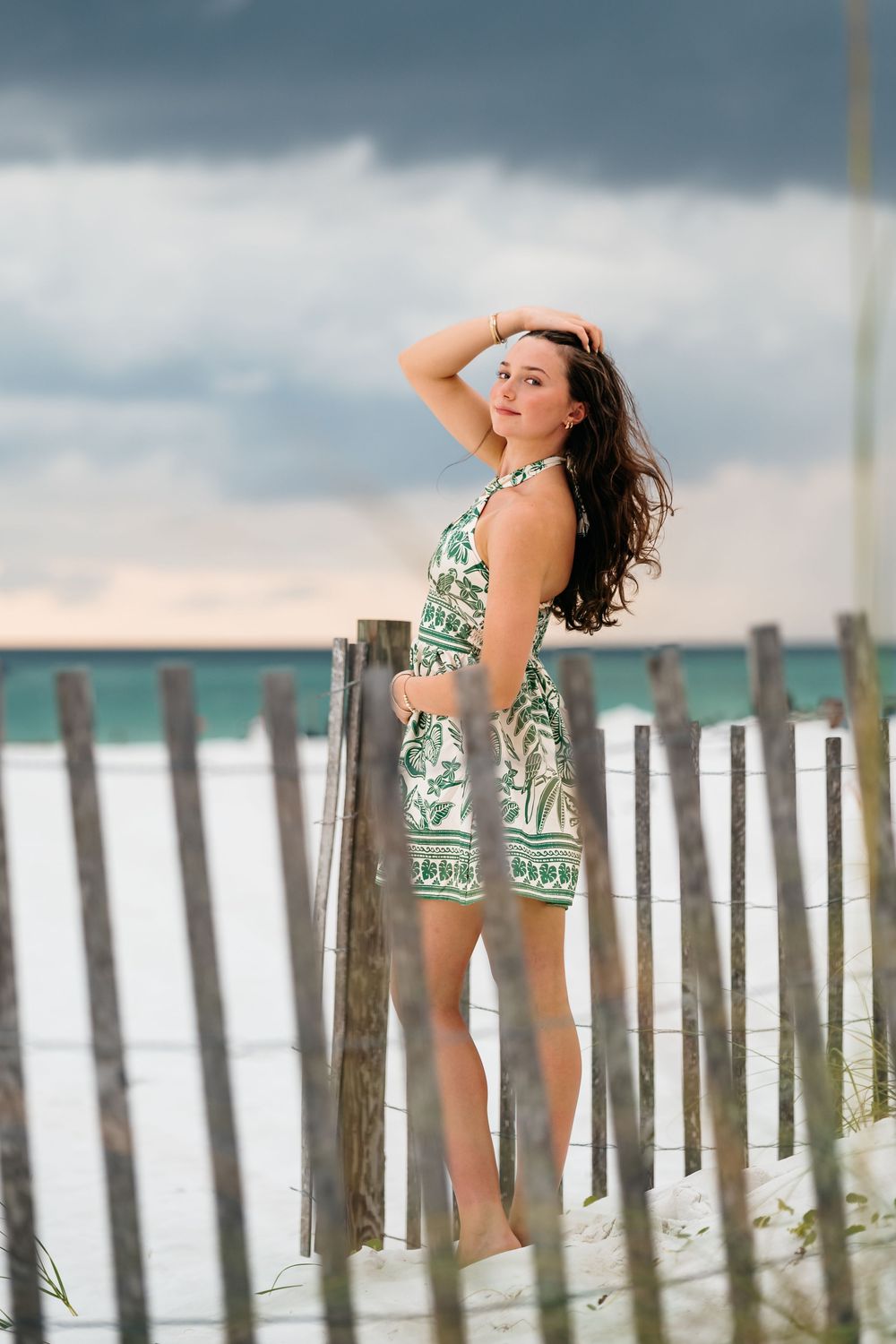 A woman in a tropical print dress poses gracefully against beach fence posts on a sandy shore.
