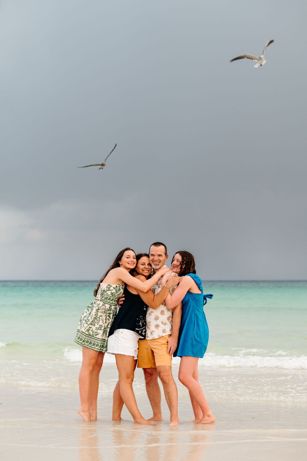 Friends share a joyful moment hugging on a tropical beach with seabirds flying overhead in a cloudy sky.