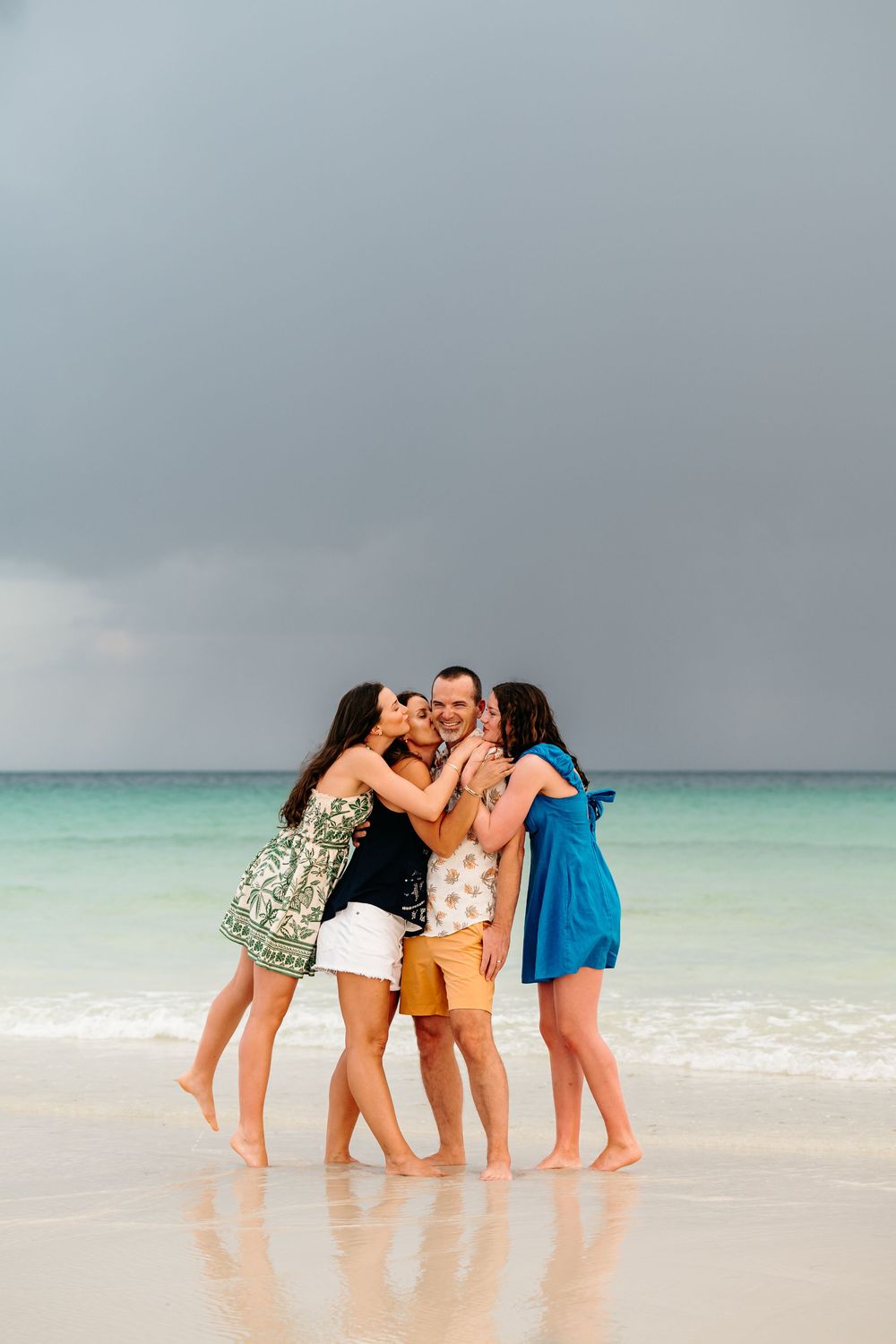 A group of friends embrace and kiss while standing in shallow turquoise ocean waters at the beach.