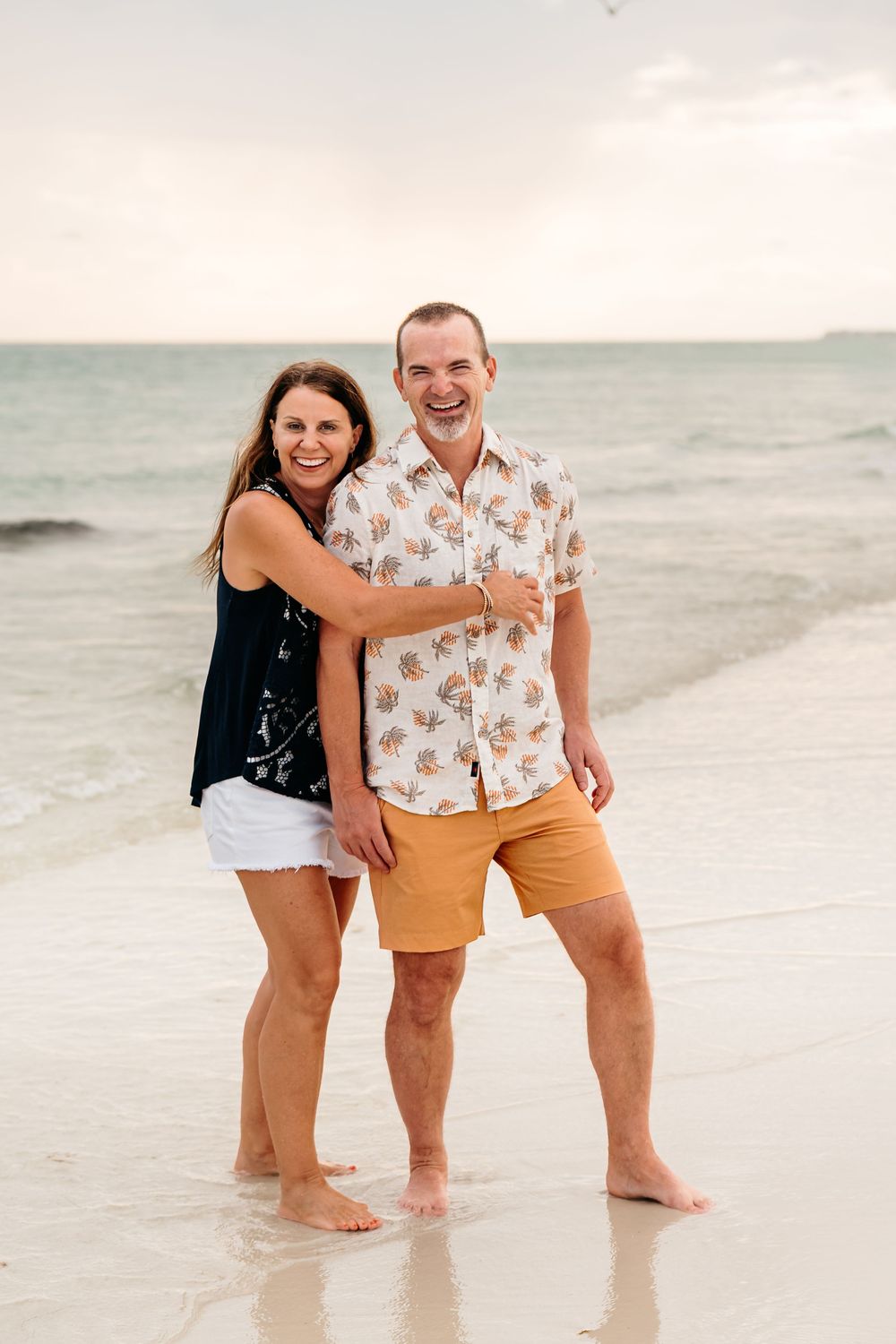 People laugh and play together in the shallow waters along a white sandy beach at sunset.