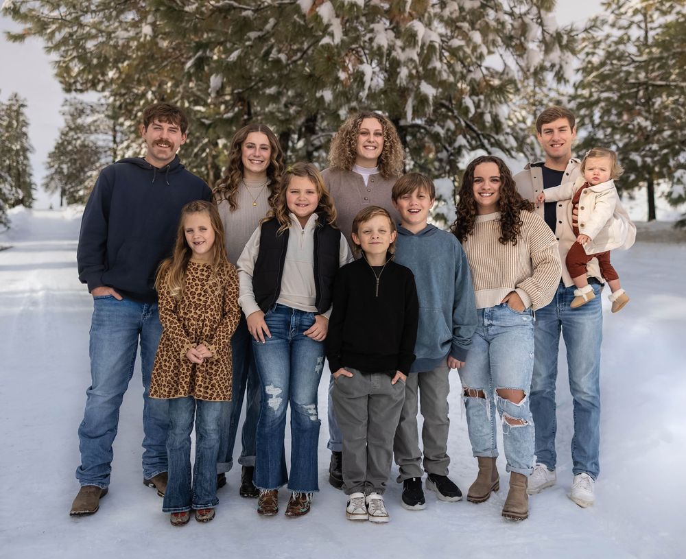 A group poses for a winter family photo sitting in snow with pine trees in the background.