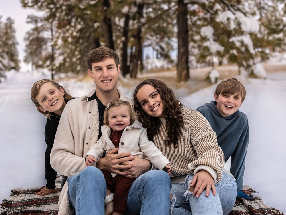 A family group sits together on a snowy ground with pine trees in the background.
