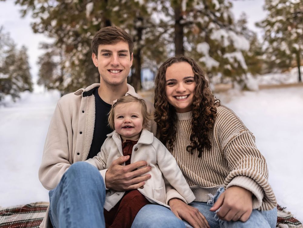 Family wearing coordinated neutral sweaters poses together in snowy winter forest setting.