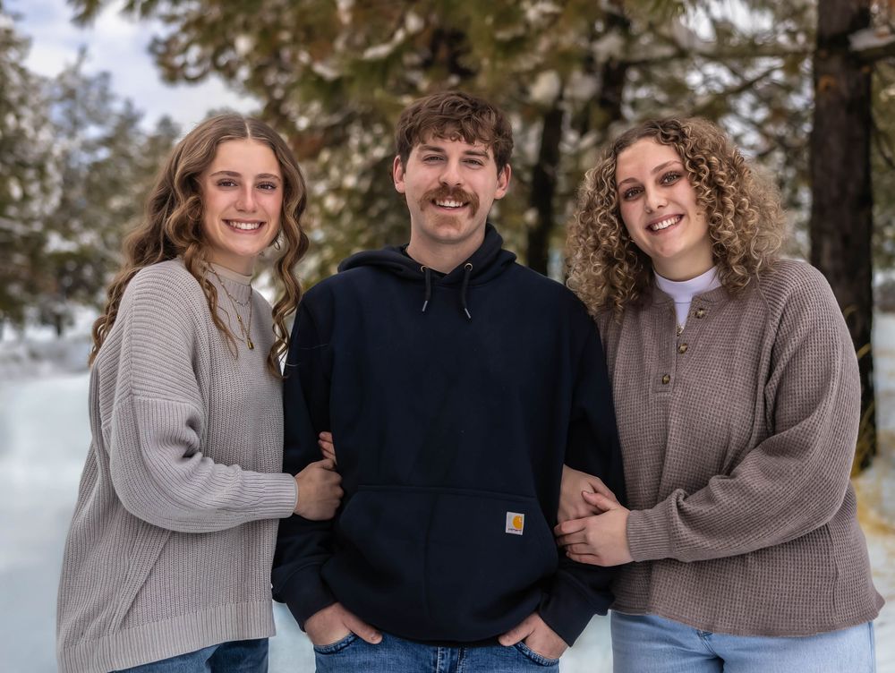 Group embraces while wearing cozy sweaters against snowy evergreen forest backdrop.