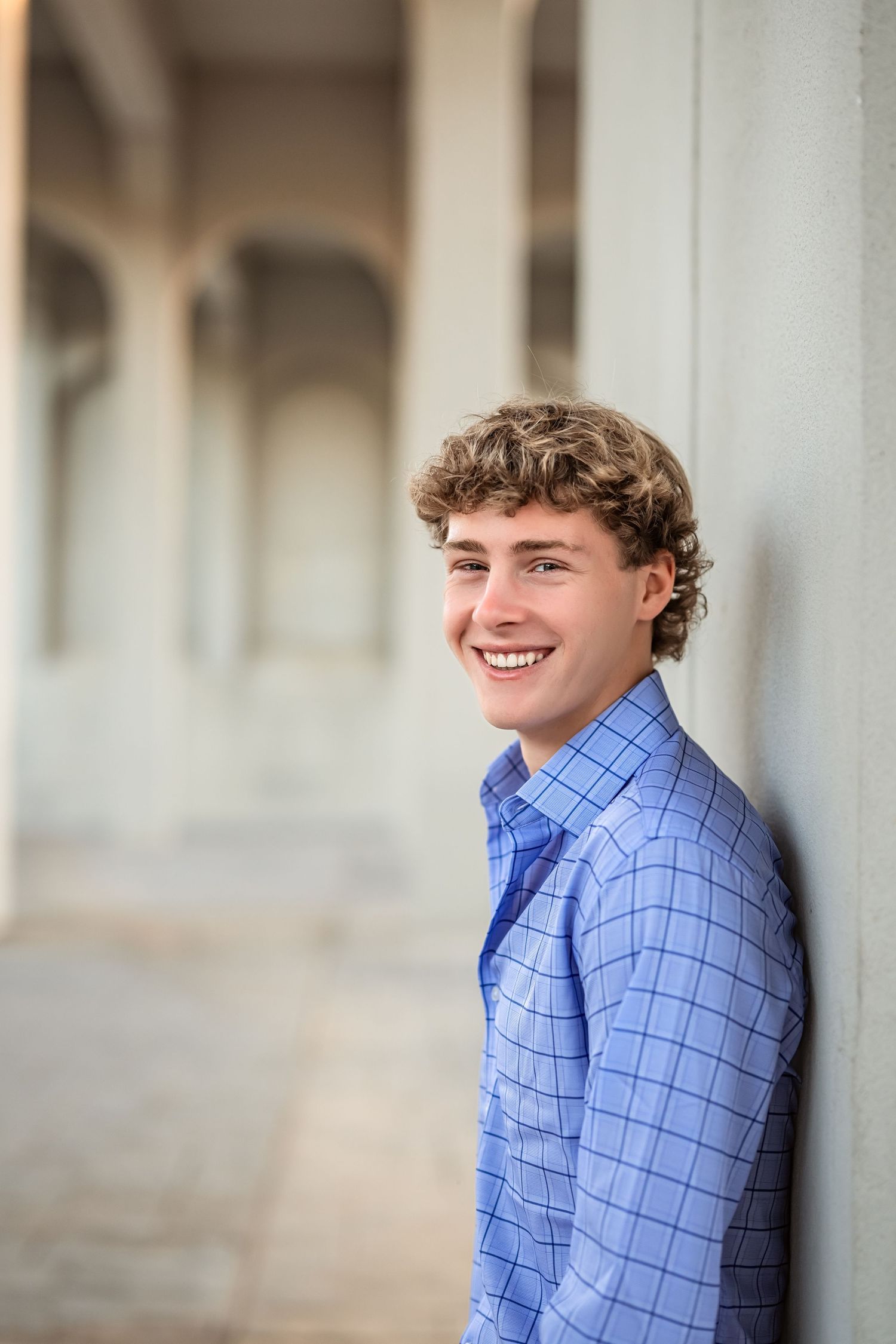 Senior boy smiles a light blue checkered dress shirt while posing against a light-colored wall in downtown Chattanooga.