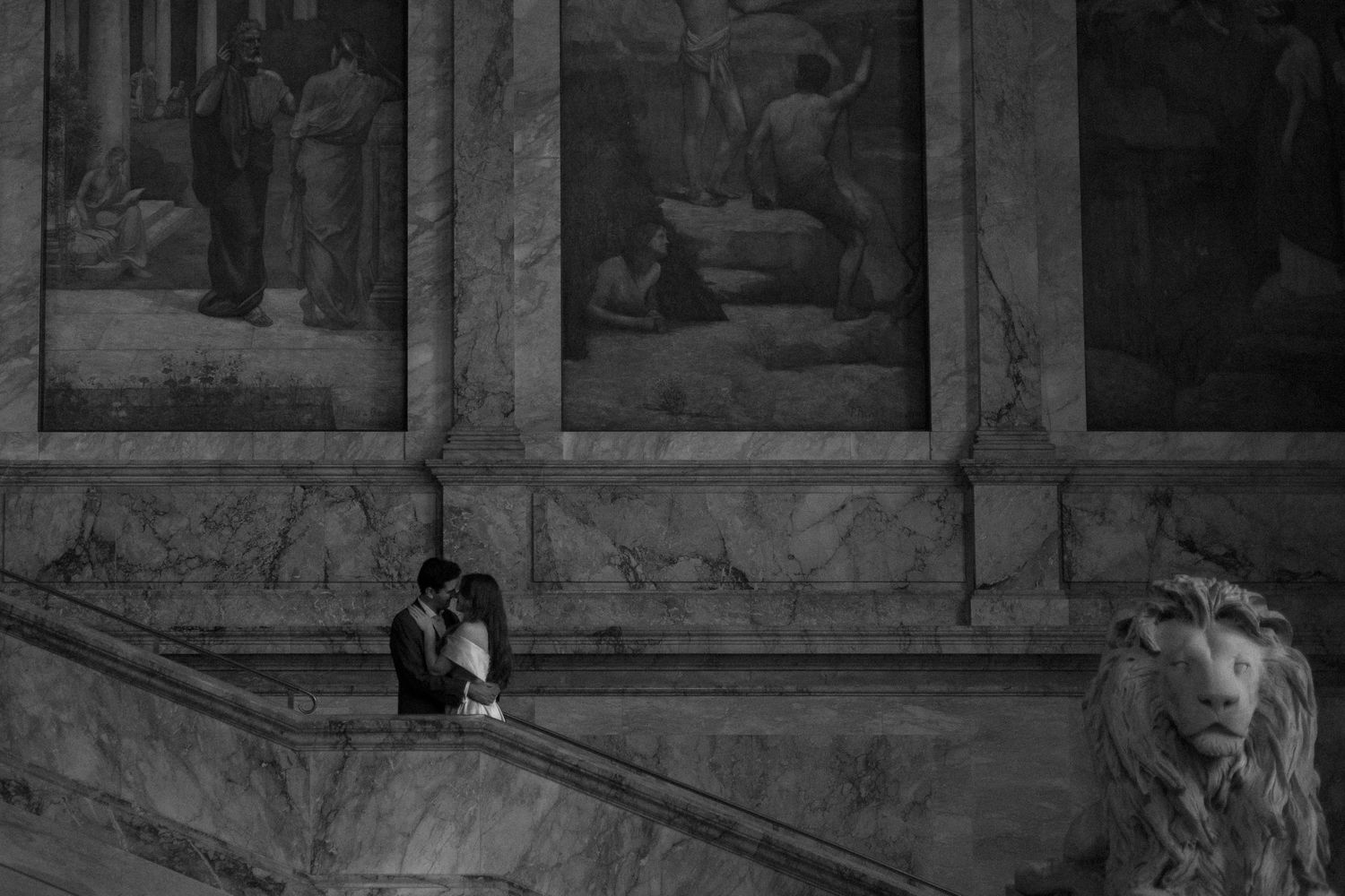 A romantic black and white scene showing two people embracing on marble steps beneath framed artwork.