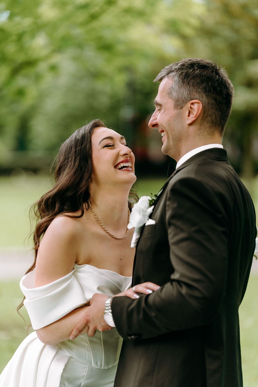 A joyful wedding couple shares a candid laughing moment together outdoors in a garden setting.