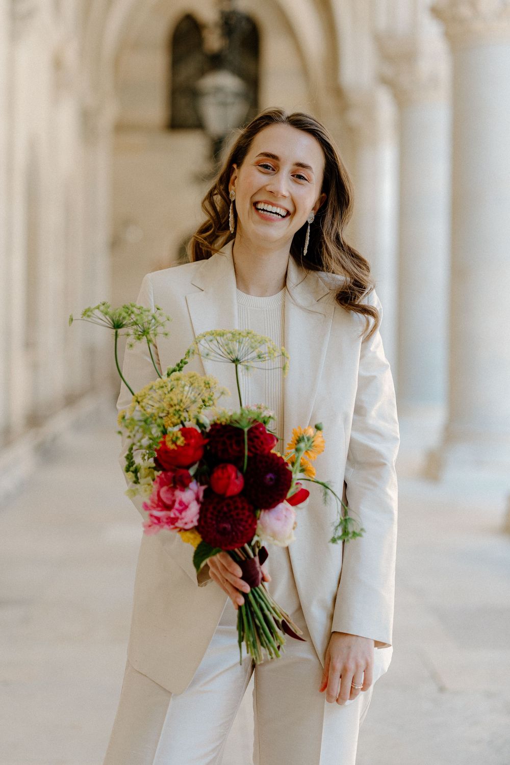 Joy radiates in this elegant white suit portrait holding a vibrant bouquet of red and pink flowers in an arched hallway.