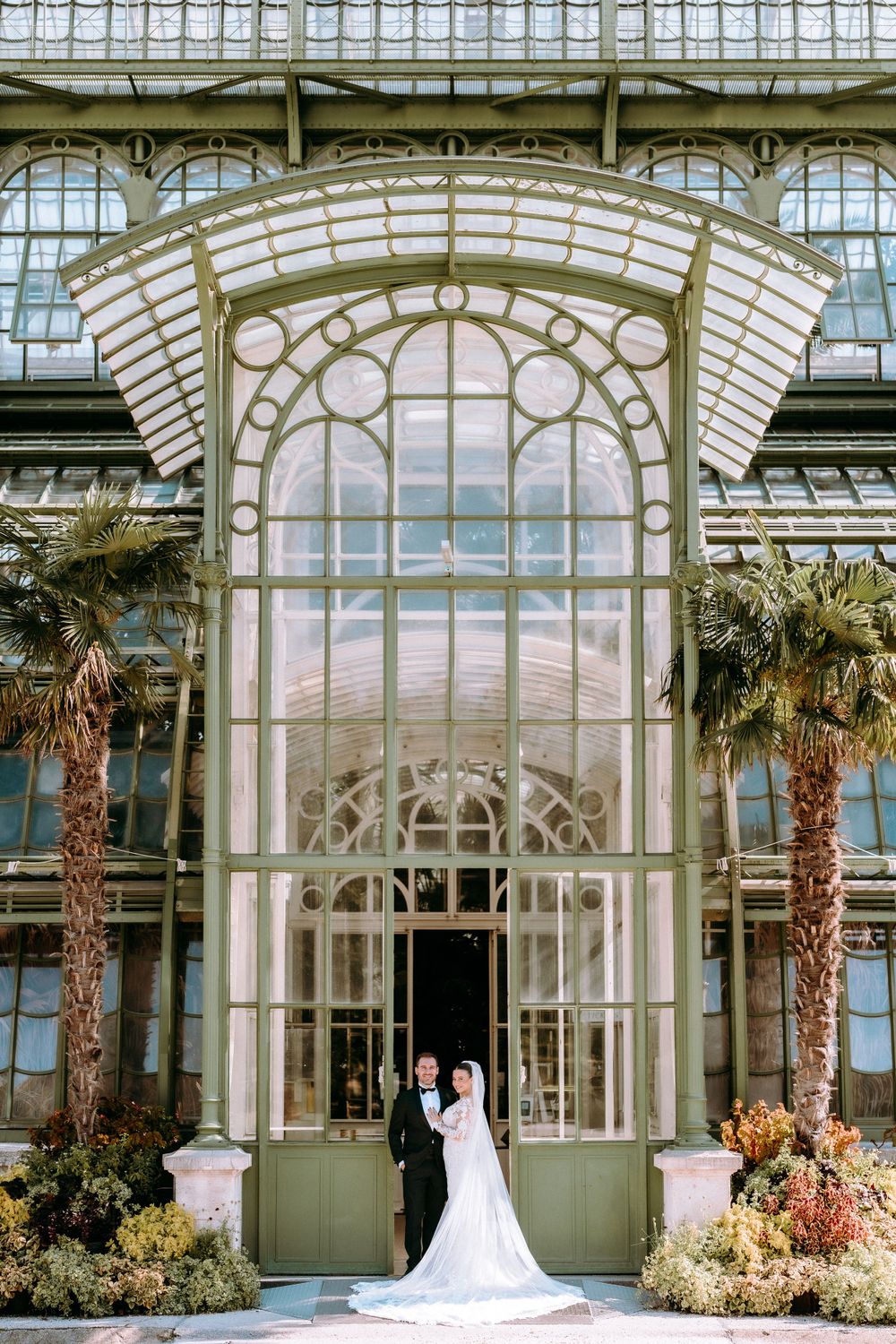 A couple stands in front of an elegant glass conservatory entrance with palm trees and ornate architectural details.