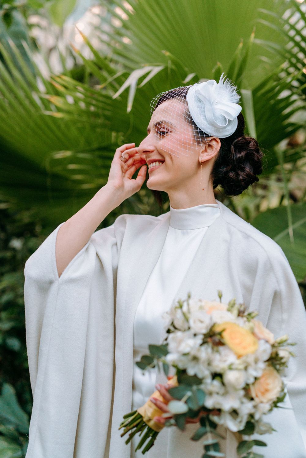 Joyful wedding moment captured among palm fronds with white dress and floral bouquet.