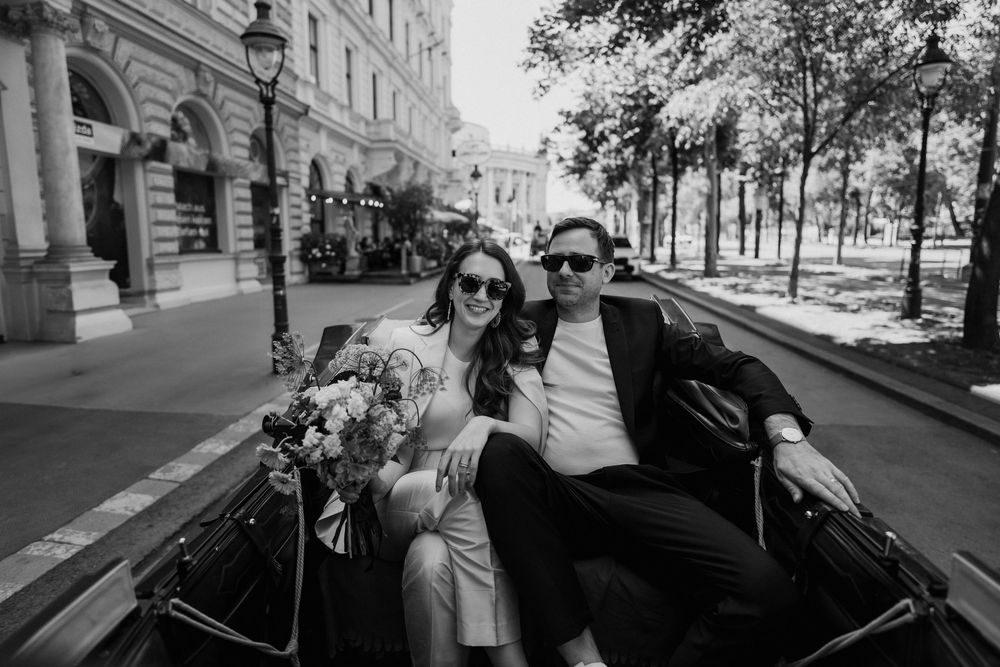 Stylish couple sits in a vintage car on a tree-lined street, with a bouquet of flowers in a black and white photograph.
