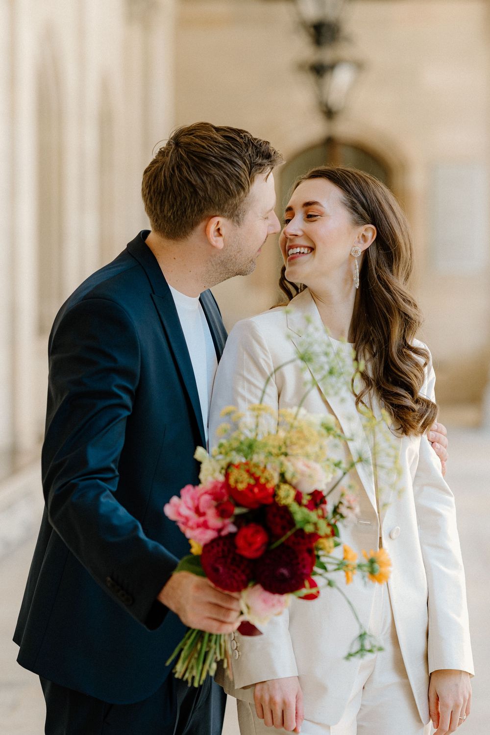A romantic couple shares an intimate moment in formal attire, with the groom holding a vibrant bouquet of flowers.