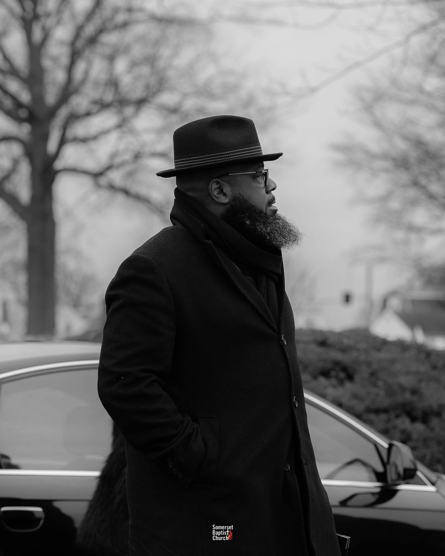 A bearded figure in a black coat and hat stands beside a car on a misty winter day.