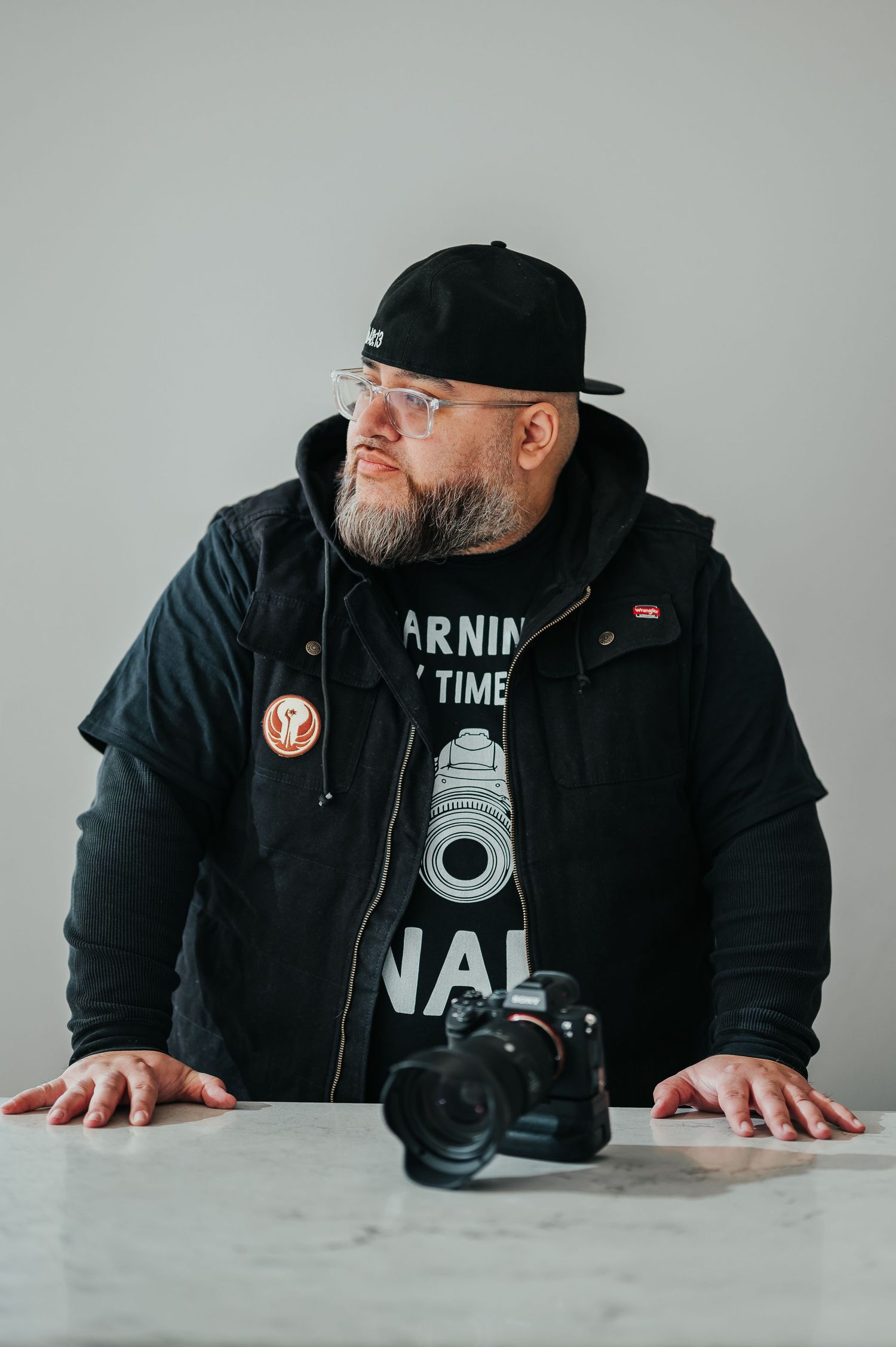 Photographer in black hoodie and cap leans over table with camera, wearing graphic t-shirt against white background.