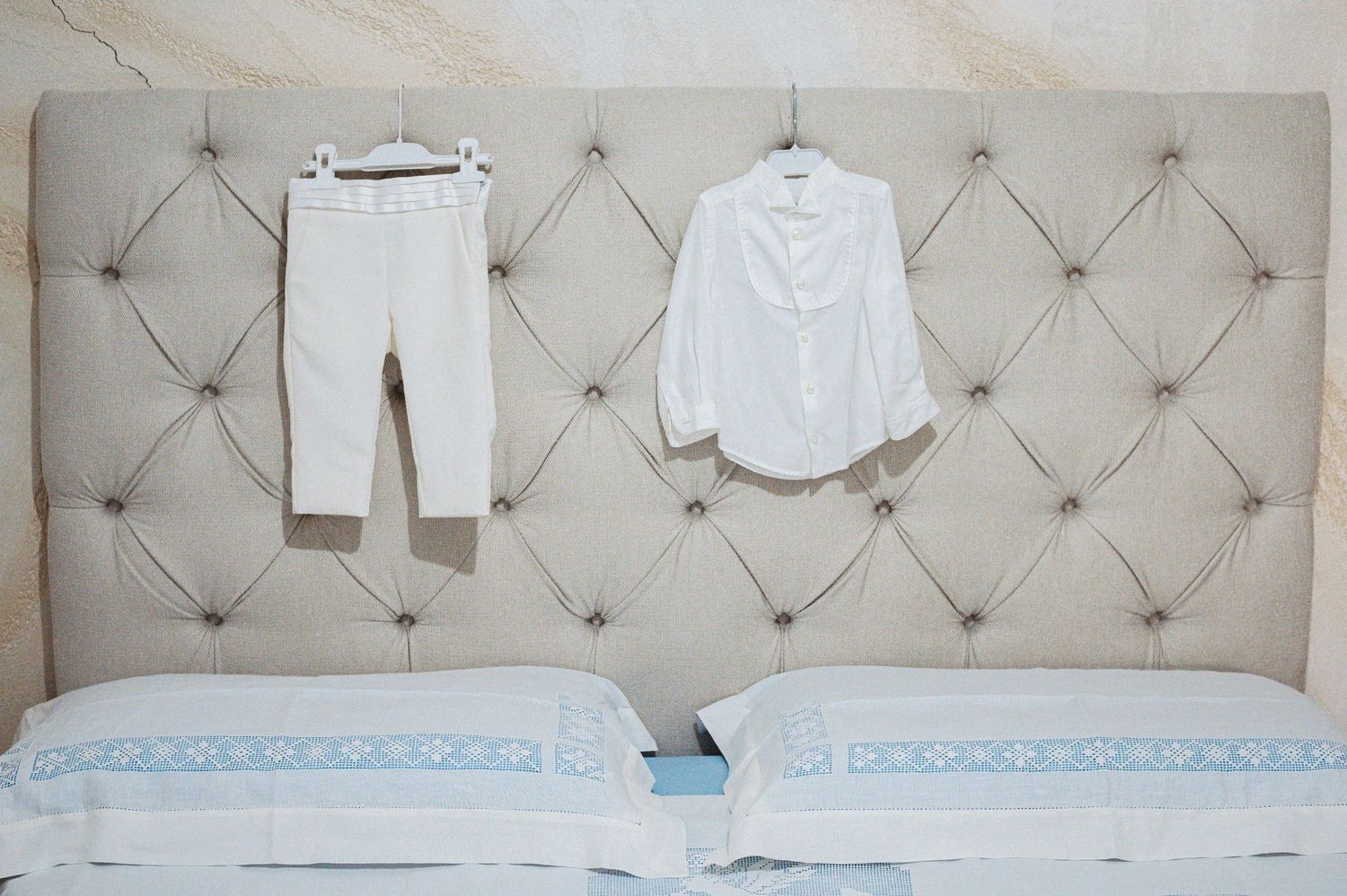 White shirt and pants hanging on a tufted beige headboard above two pillows.