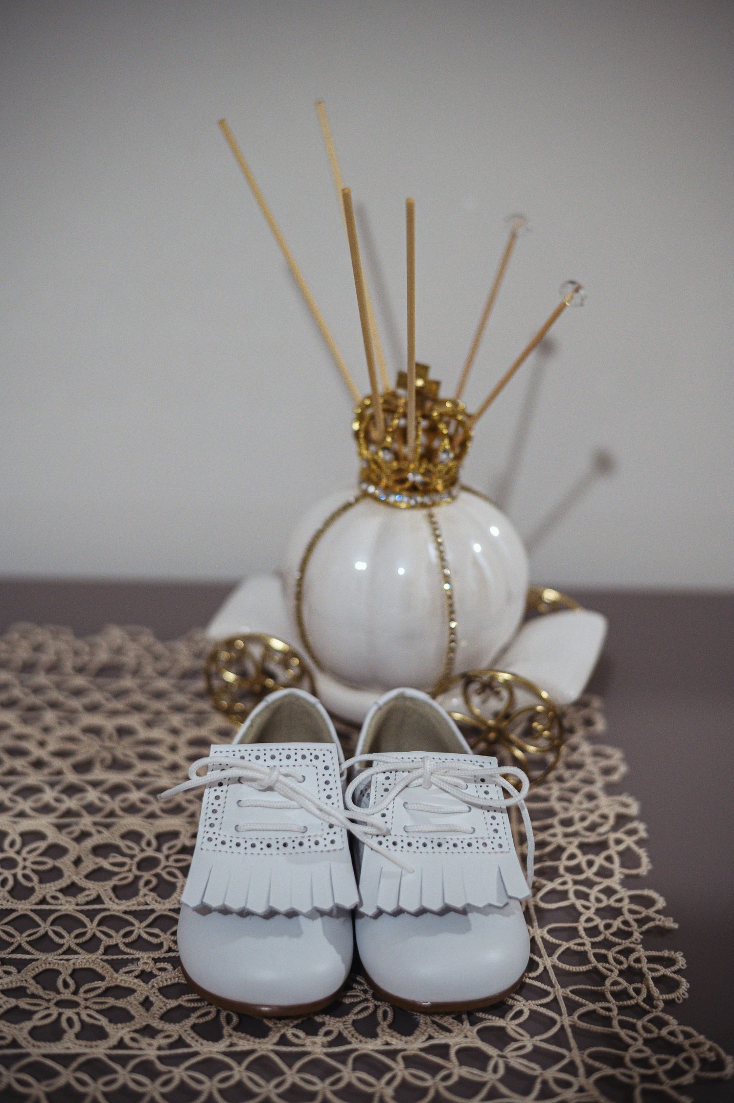 White baby shoes with fringe detail sit next to a decorative crown vase on lace doily.
