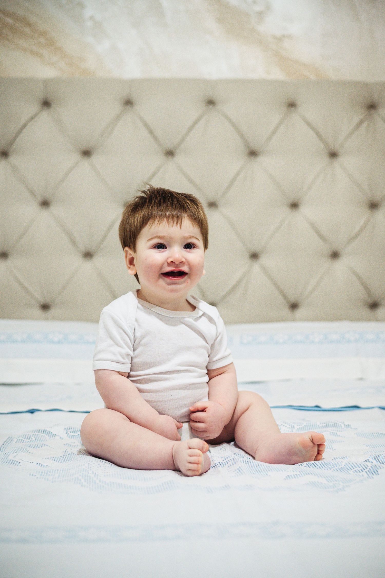 Baby in white onesie sits happily on blue bedding against tufted headboard.