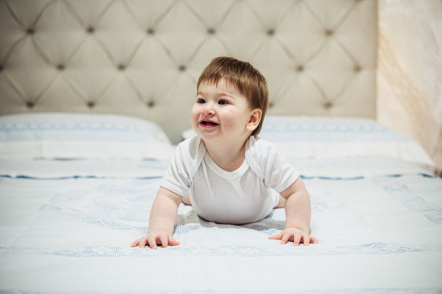 Smiling baby in white onesie crawls on blue bedding.