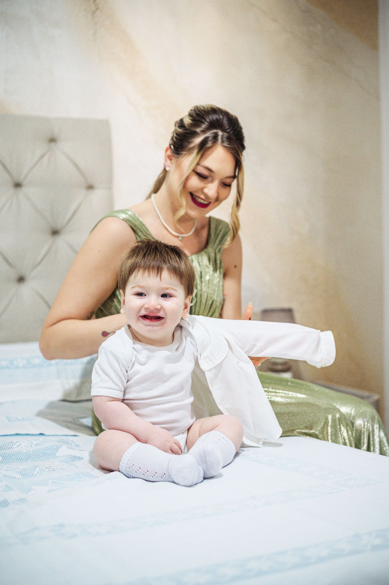 Joyful moment shared between caregiver and baby in white onesie on bed.
