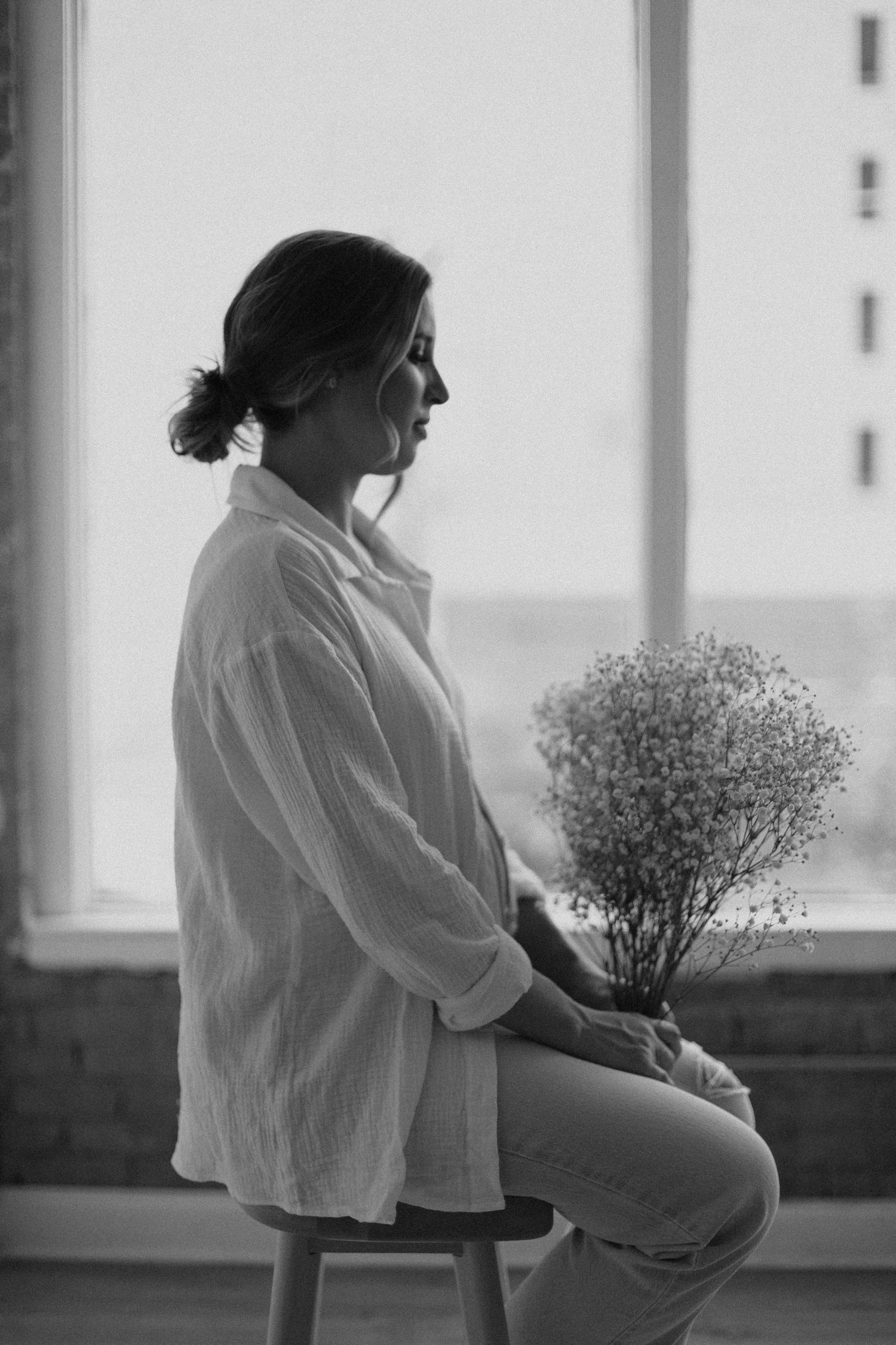 Black and white portrait of person holding baby's breath flowers while sitting by window.