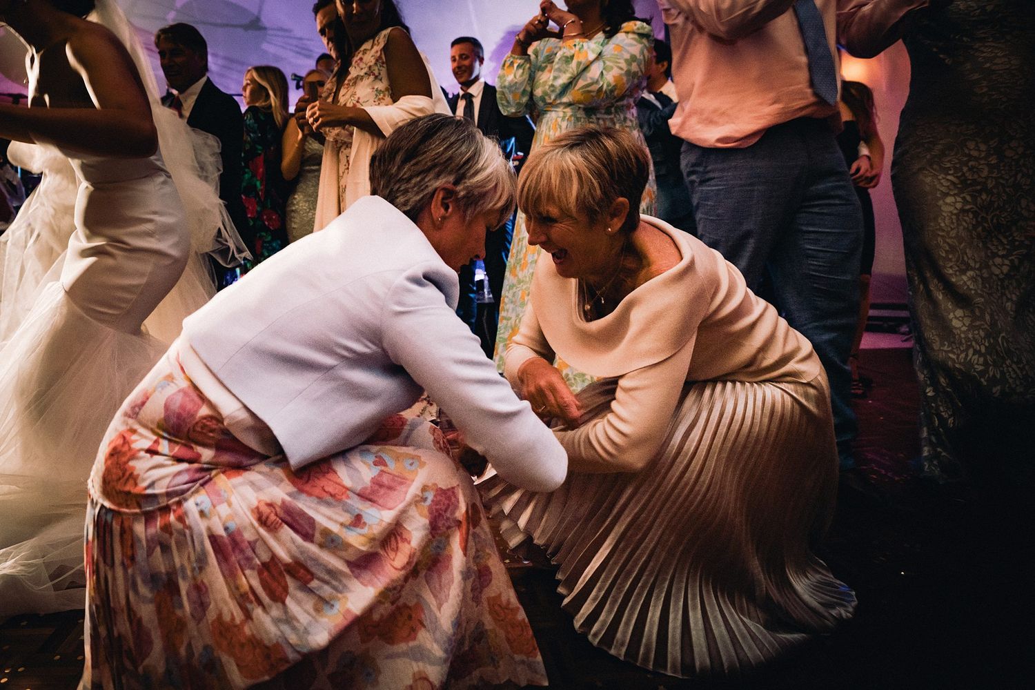 Two elegantly dressed guests in floral and pleated skirts embrace while dancing at an evening wedding at Fox Harb'r Resort wedding venue.