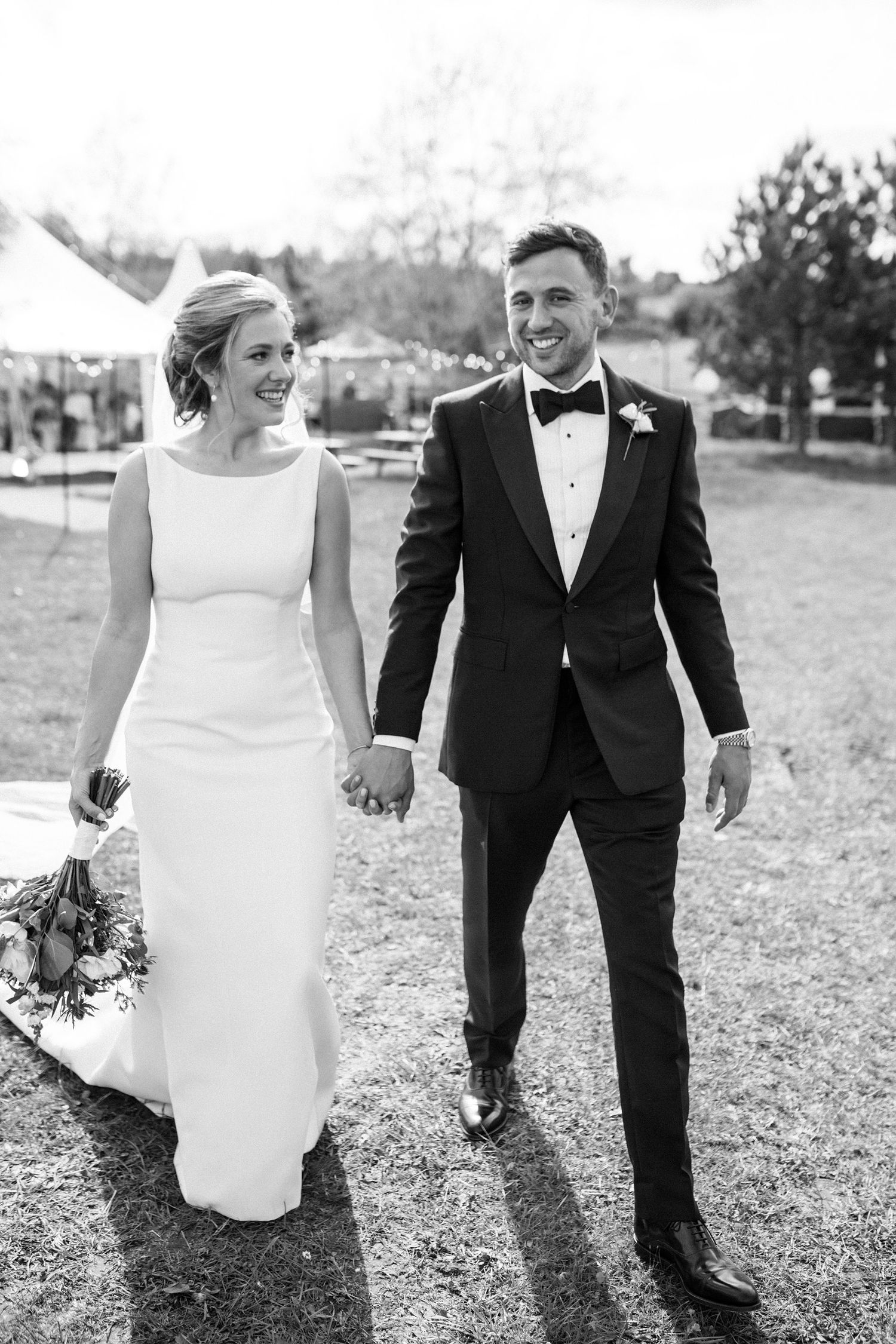 A black and white wedding photograph of a couple walking outdoors at Fox Harb'r Resort, with the bride in a sleek gown and groom in tuxedo