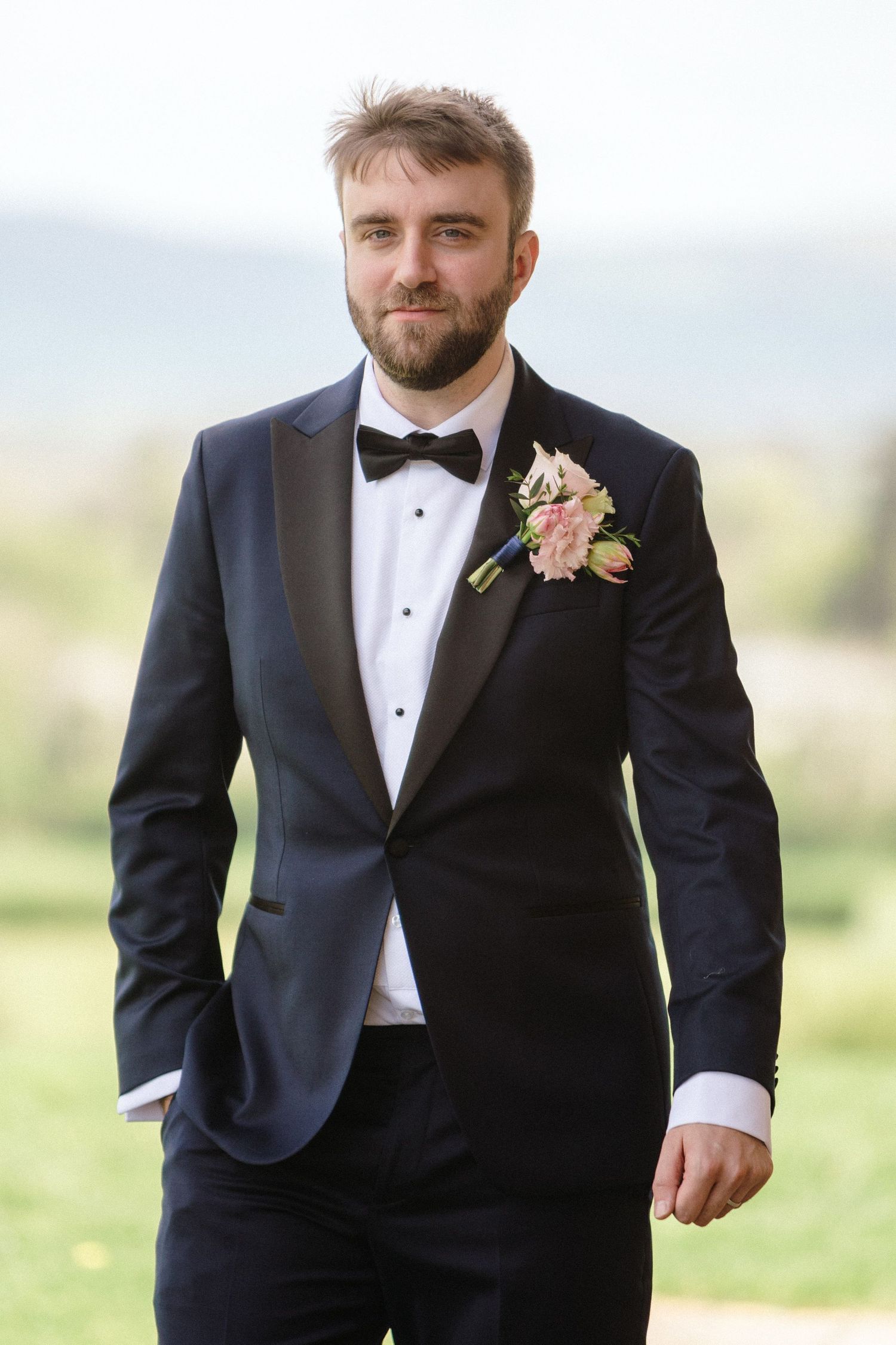 Groom in classic black tuxedo with bow tie and peach floral boutonniere poses outdoors against blurred landscape at Fox Harb'r Resort