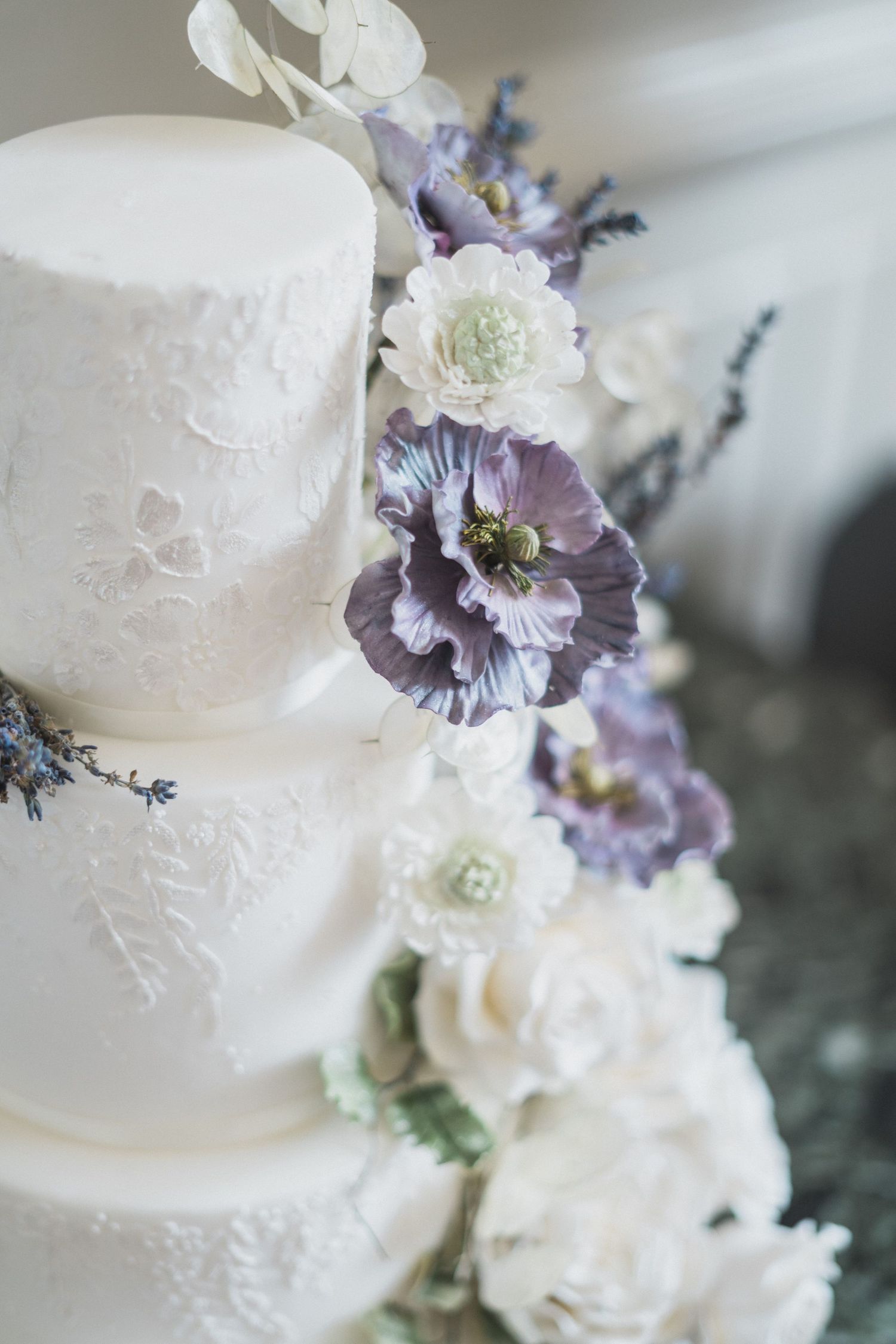 Elegant white wedding cake decorated with delicate purple and white floral arrangements and lace details.