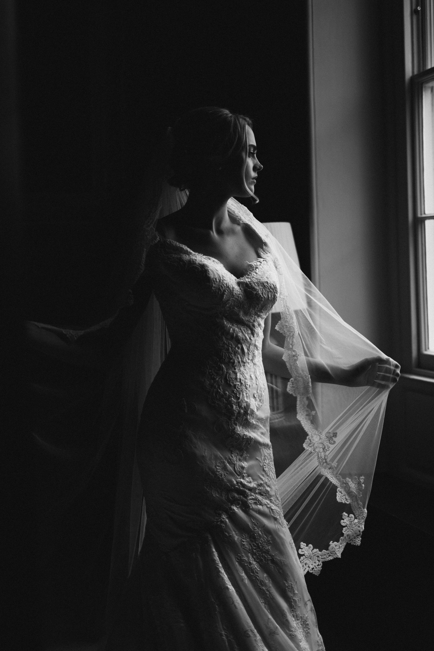 Black and white portrait of a bride in a lace wedding gown standing by window with flowing veil in dramatic lighting at Fox Harb'r Resort