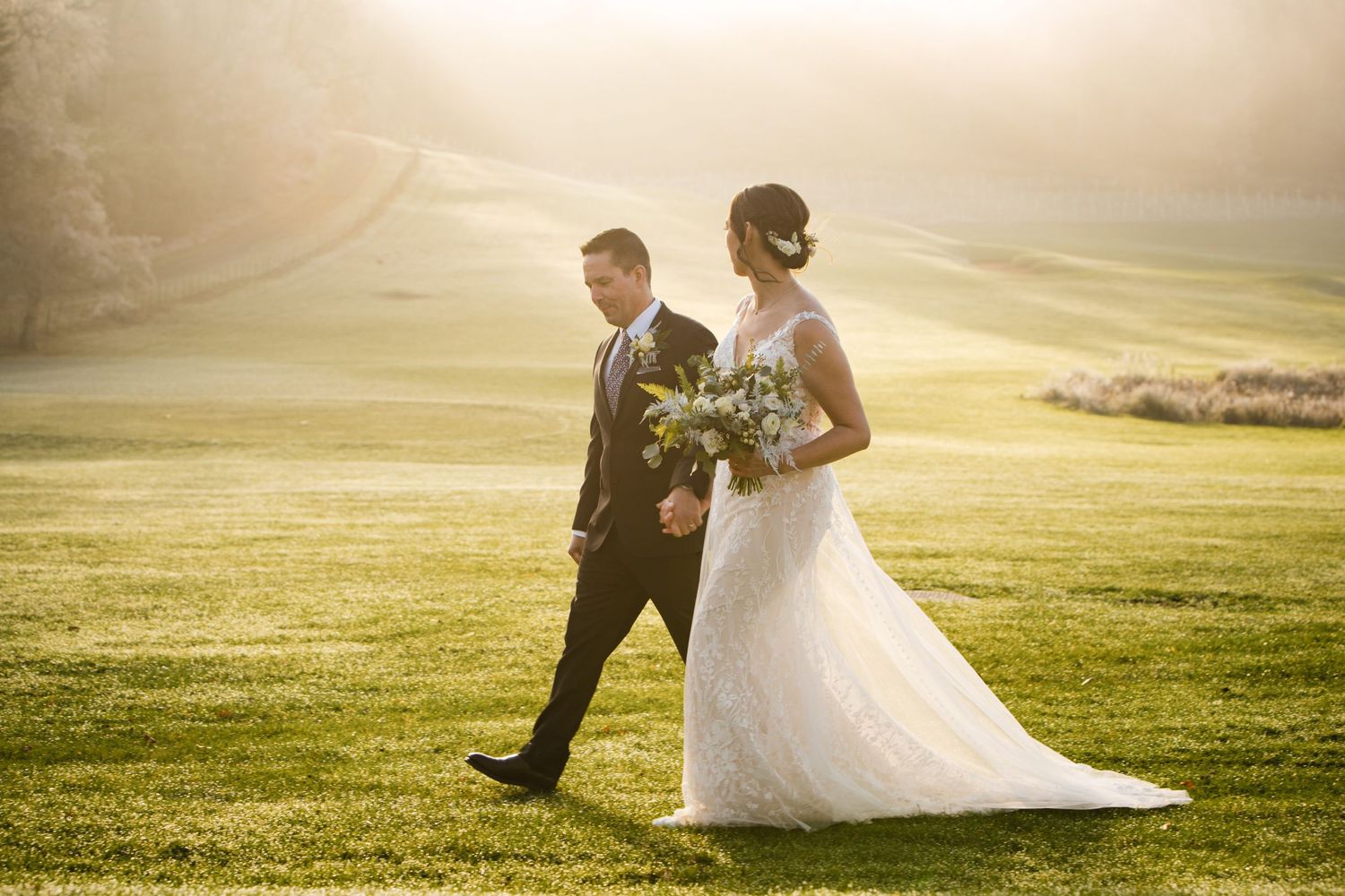A bride and groom walk together across a sunlit golf course during golden hour at Fox Harb'r Resort with her white gown trailing behind.