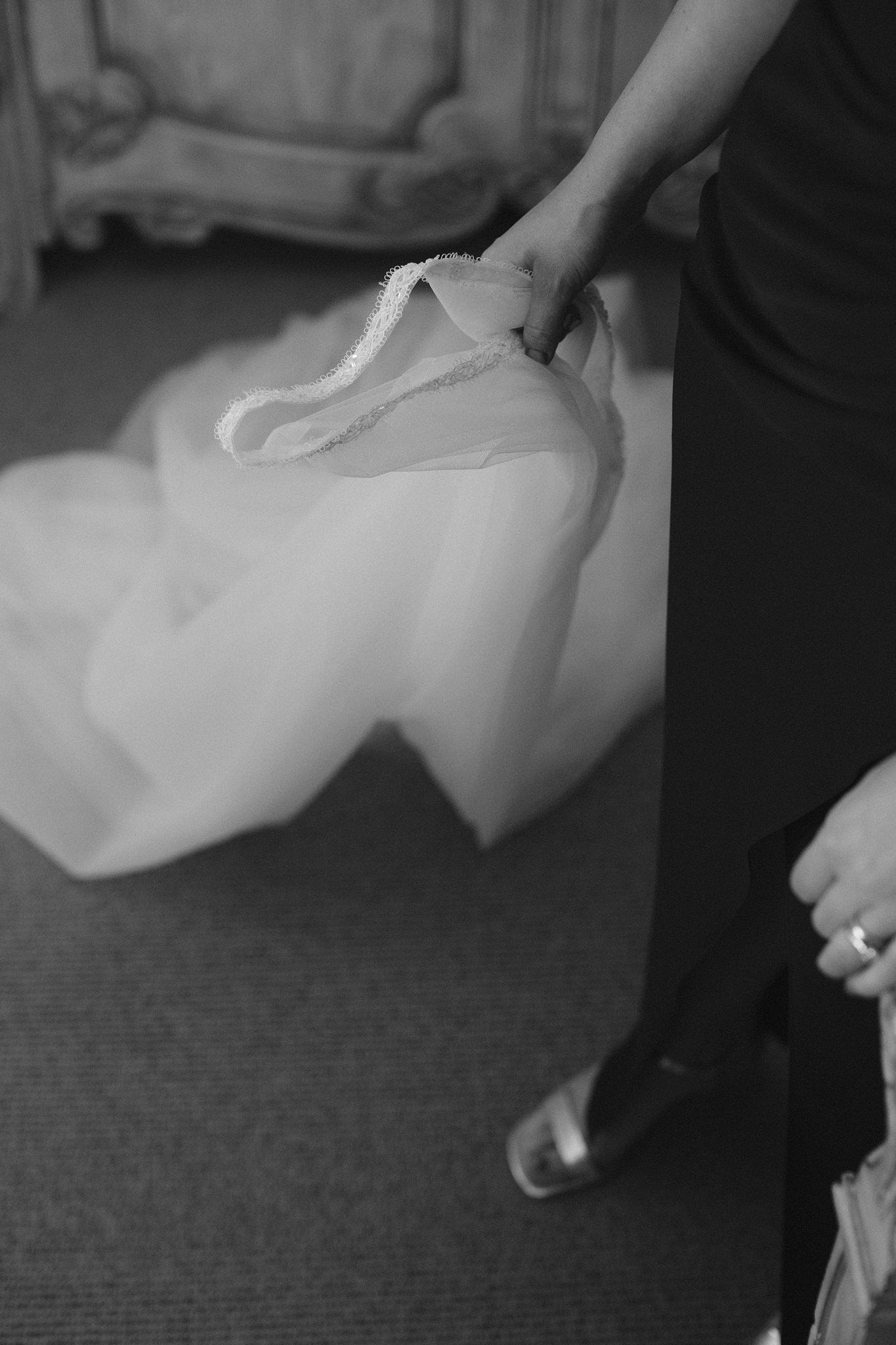 Black and white close-up shot of hands holding a flowing white wedding dress train against carpeted floor at Fox Harb'r Resort