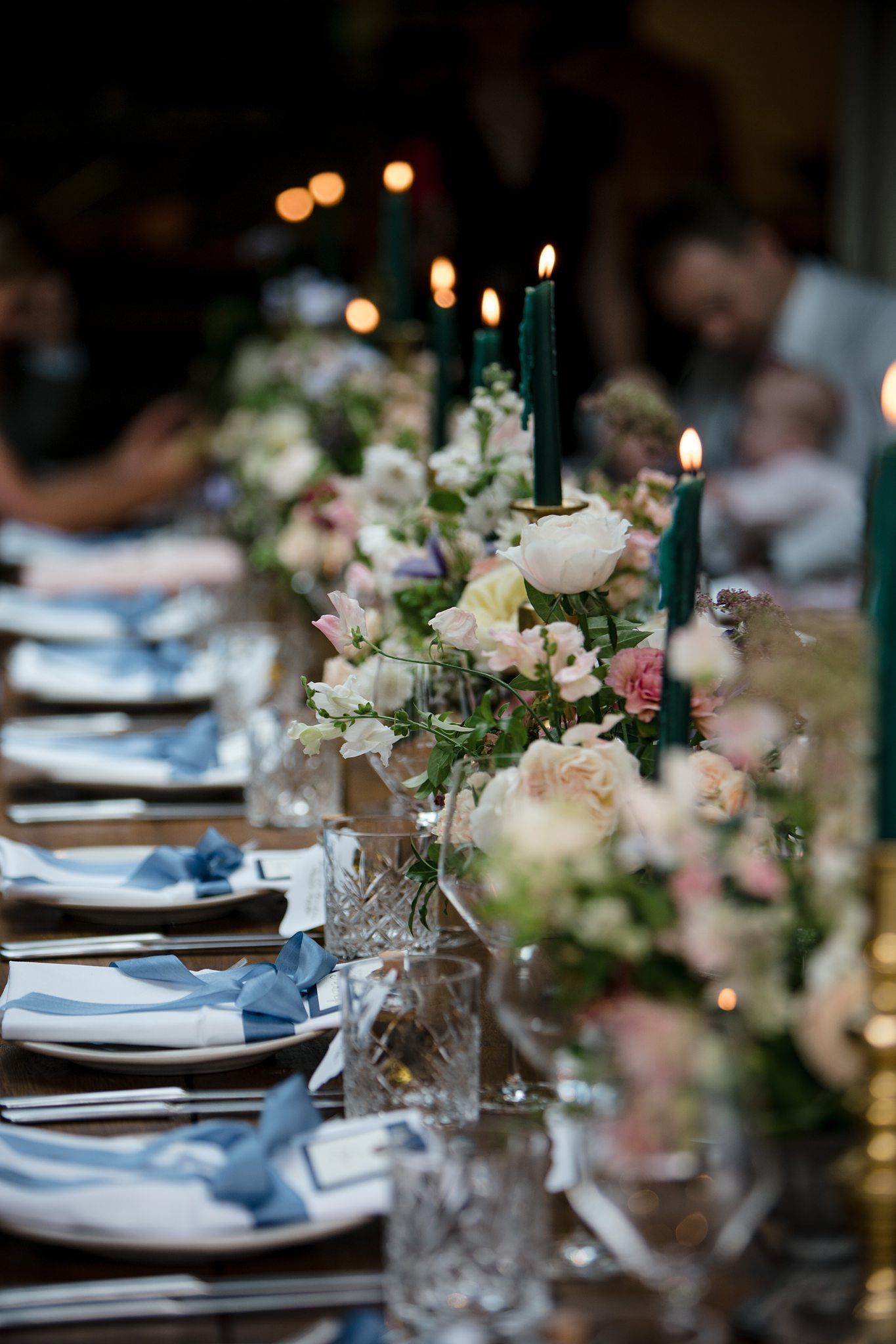 Elegant wedding table setting with blue napkins, tall green candles, and soft pink and white floral centerpieces at Fox Harb'r wedding