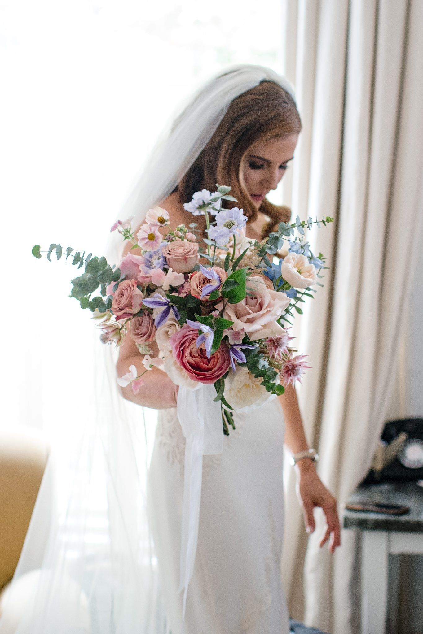 A bride in white wedding dress holds an elegant bouquet of pink and blue flowers with eucalyptus against natural light Fox Harb'r Resort.