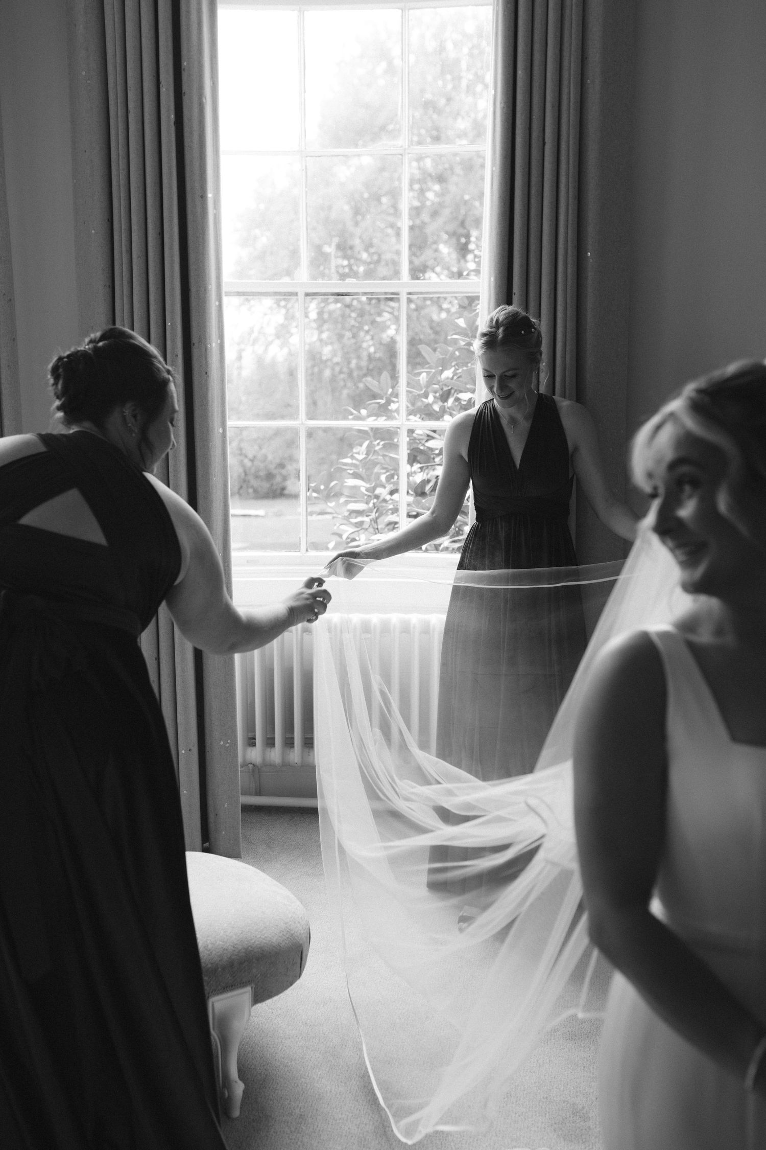 A black and white photograph shows bridesmaids helping adjust a bride's flowing veil near a bright window at Fox Harb'r Resort wedding venue