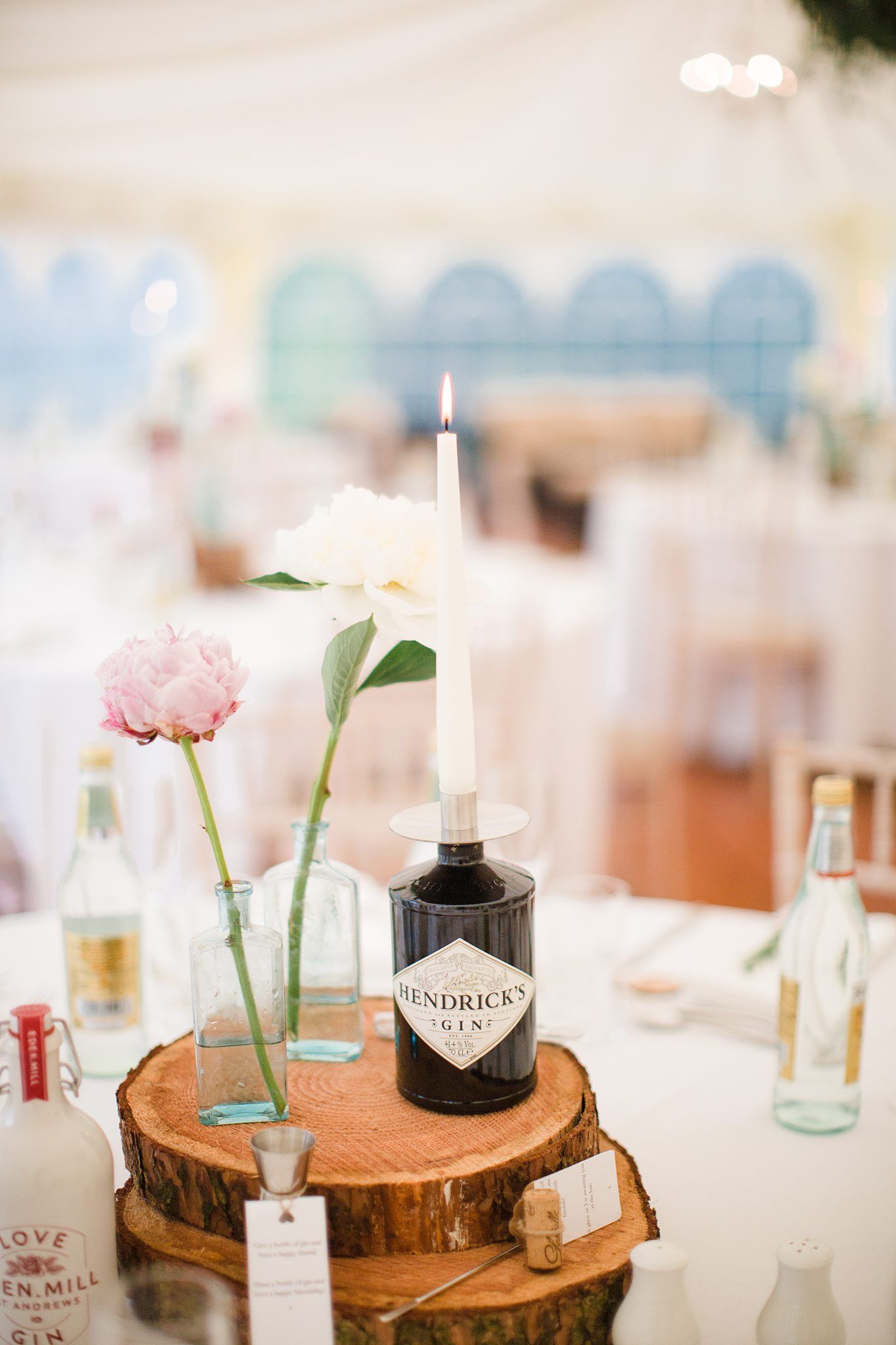 Rustic wedding centerpiece with wooden log slices, pink peony in glass vase, and black Hendrick's gin bottle with white candle at Fox Harb'r
