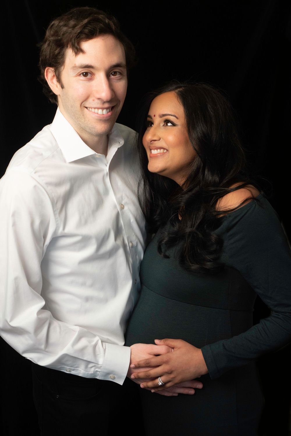 Expectant couple embracing in formal attire against dark background.