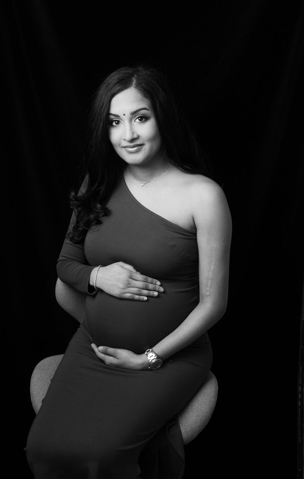 Black and white maternity portrait of woman in one-shoulder dress, looking at the camera.