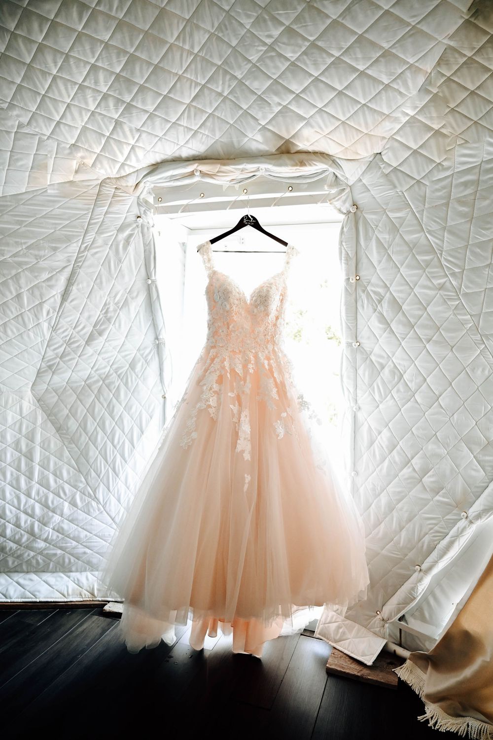 A blush pink tulle wedding gown with lace details hangs elegantly against a white quilted background.