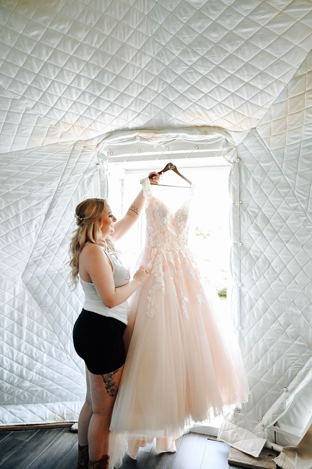 A blush pink tulle wedding dress hangs from a hanger as someone adjusts it in a bright white quilted room.