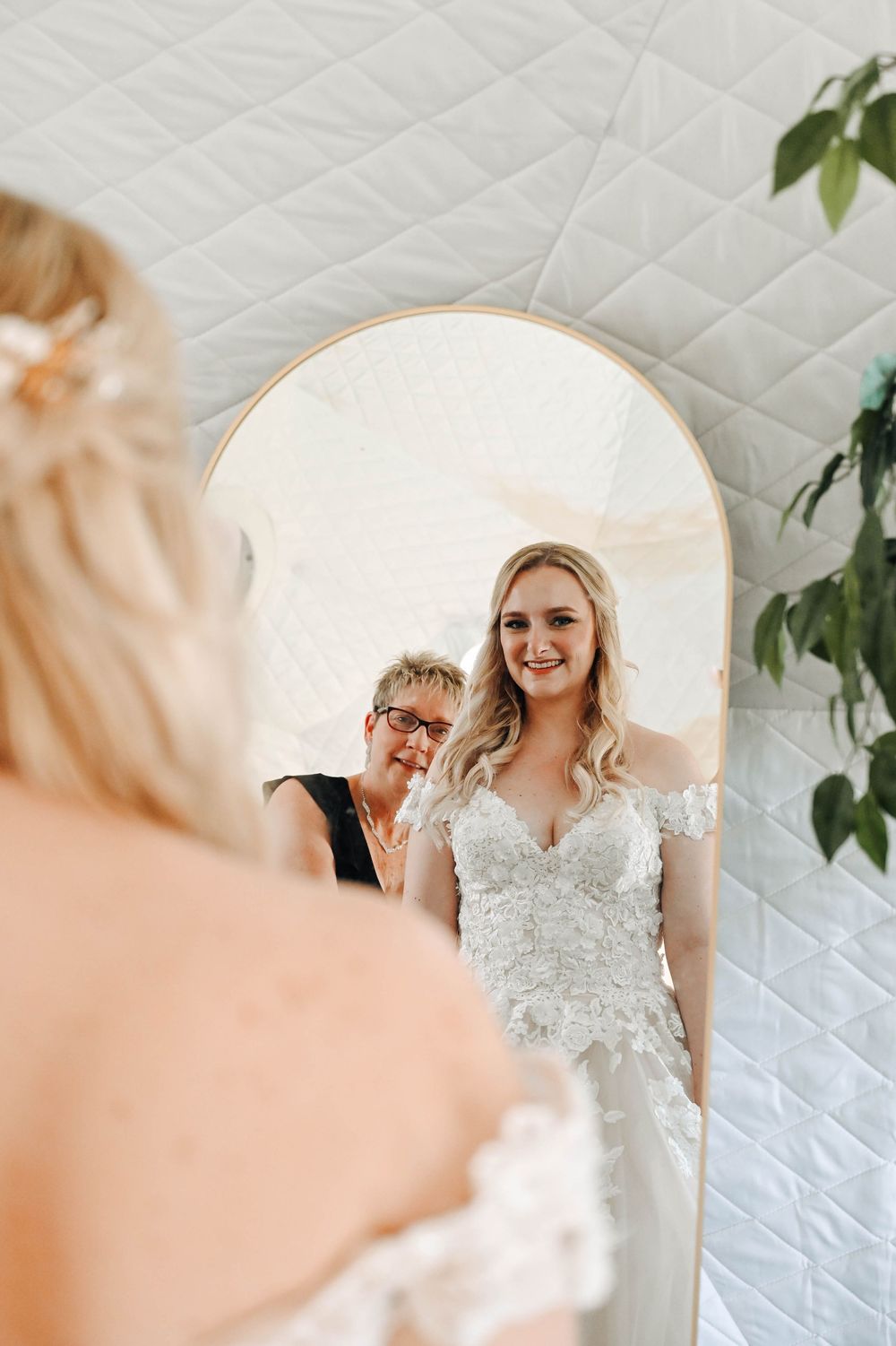 A person wearing a white wedding dress looks in a large mirror reflecting soft natural light.