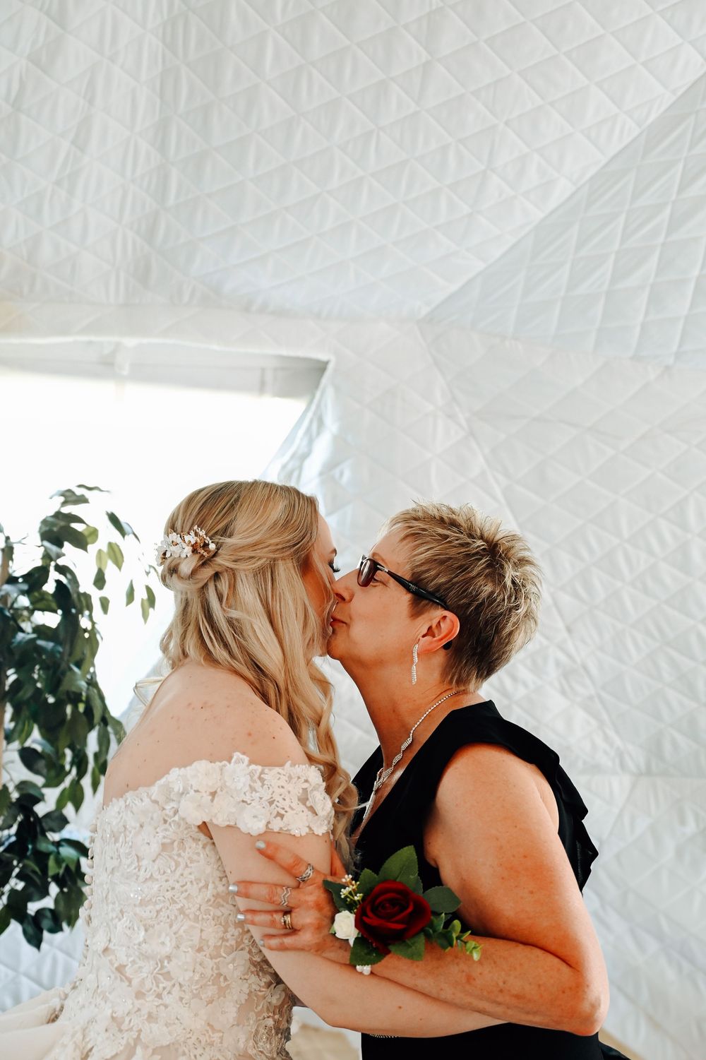Two people share a tender kiss during a wedding ceremony in a bright white venue with greenery decor.