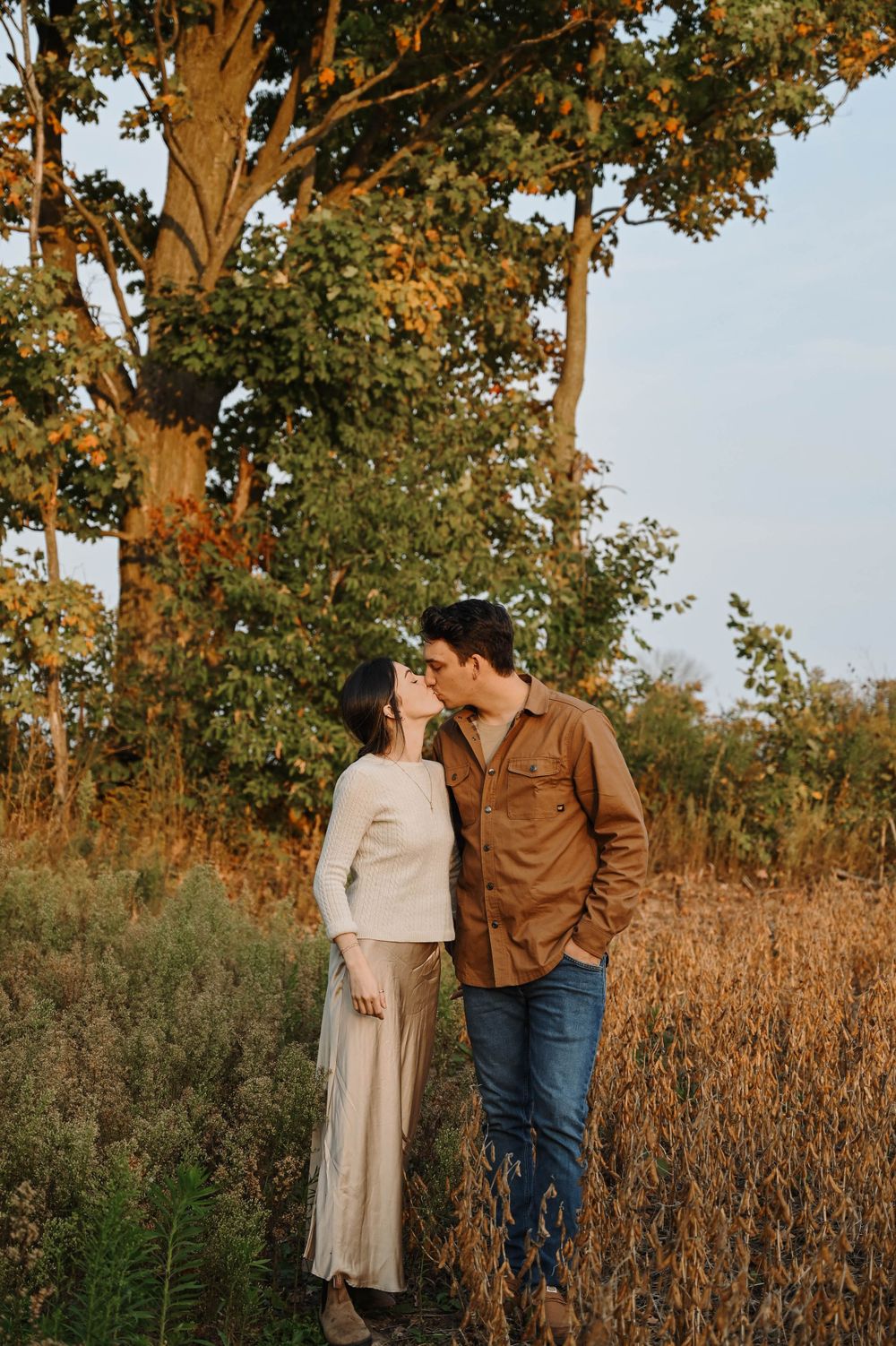 A tender romantic moment captured at sunset in a golden field with tall grass and autumn trees in the background.